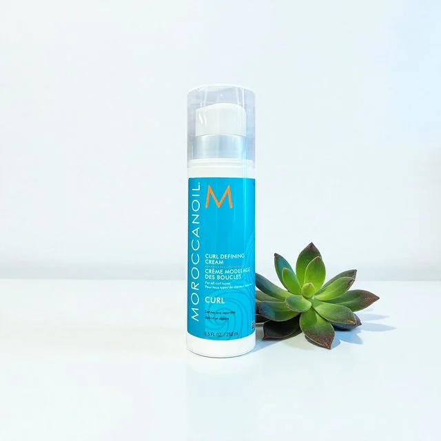 A blue and white bottle of Moroccanoil Curl Defining Cream standing next to a green succulent plant on a white surface.