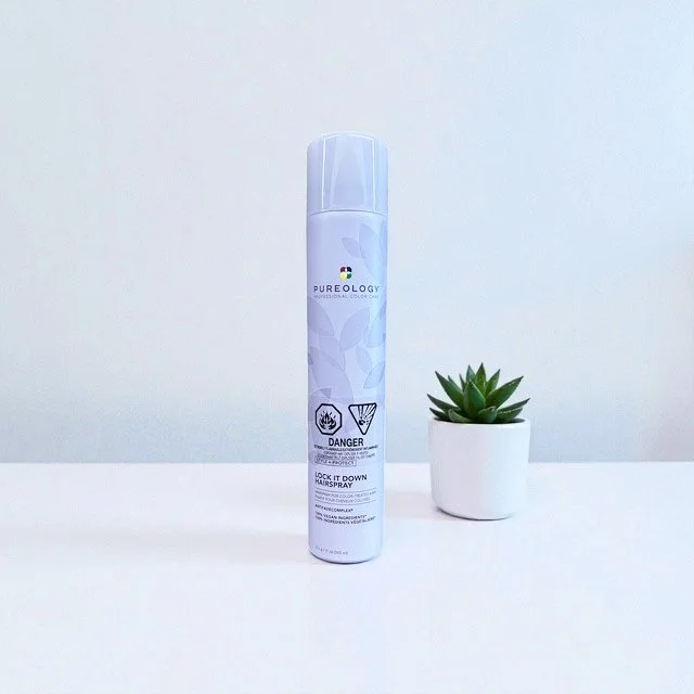 A can of Pureology Lock It Down Hairspray next to a small potted succulent plant on a white surface against a neutral background.