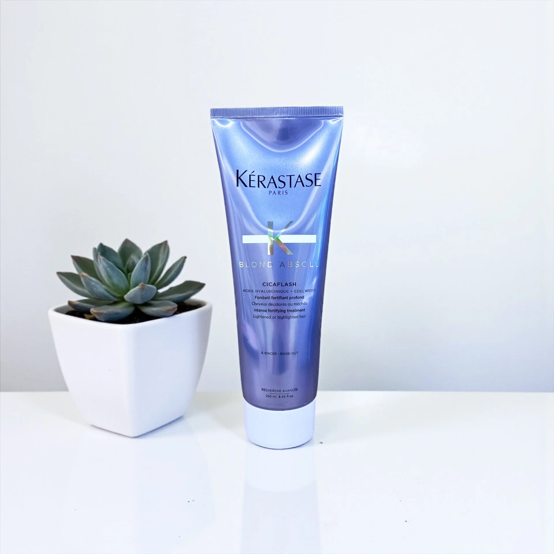 A tube of Kérastase Blond Absolu Cicaflash hair treatment standing on a white surface next to a potted succulent plant.