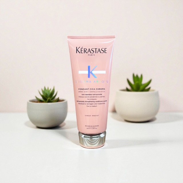 Pink tube of Kerastase Chroma Absolu Fondant Cica Chroma  hair care product positioned between two small potted succulent plants on a white surface with a light pink background.