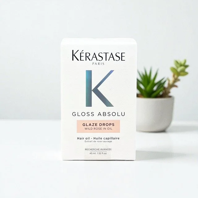 Kérastase Paris Gloss Absolu glaze drops hair oil in a white box with pink label, placed on a white surface with a small potted succulent plant in the background.