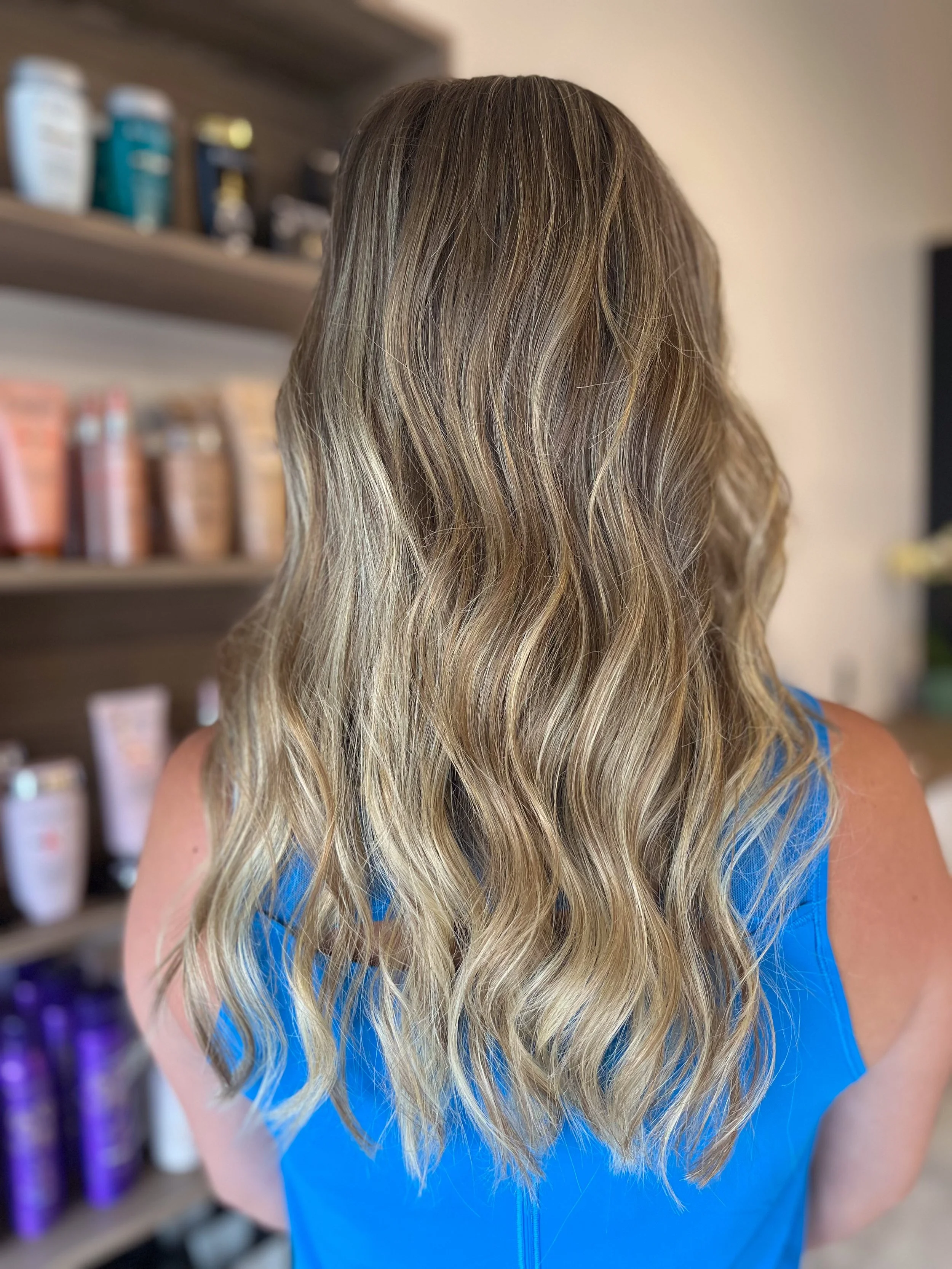Back of a woman with long, wavy blonde hair wearing a blue top, standing in front of a shelf with hair products.