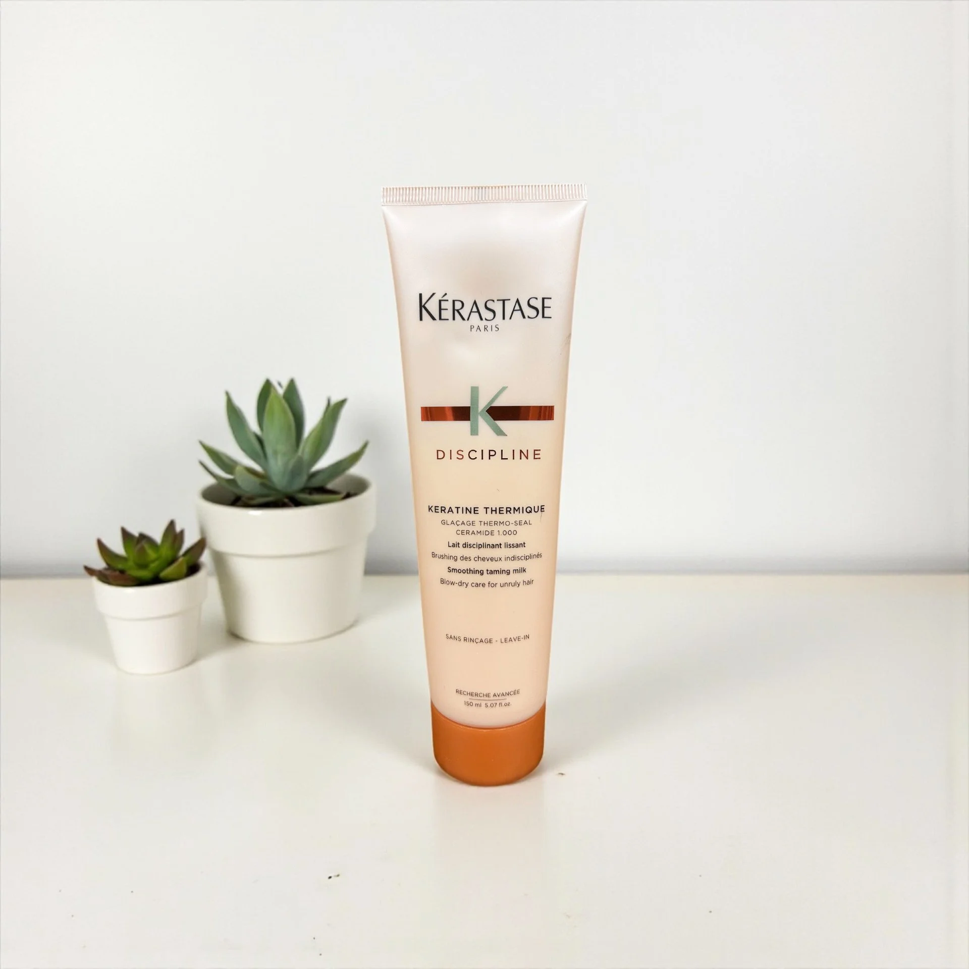 Tube of Kérastase Discipline Keratine Thermique on a white surface with two potted succulents in white pots behind it.