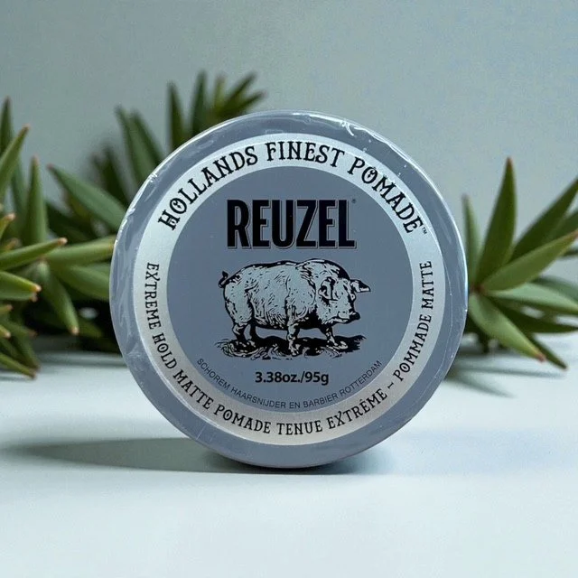 A tin of REUZEL High Shine Matte Pomade, 3.38 oz, with packaging that features a pig illustration, on a white surface with green plant leaves in the background.
