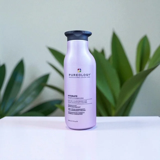 A bottle of Pureology Hydrate Shampoo on a white surface with green leaves in the background.