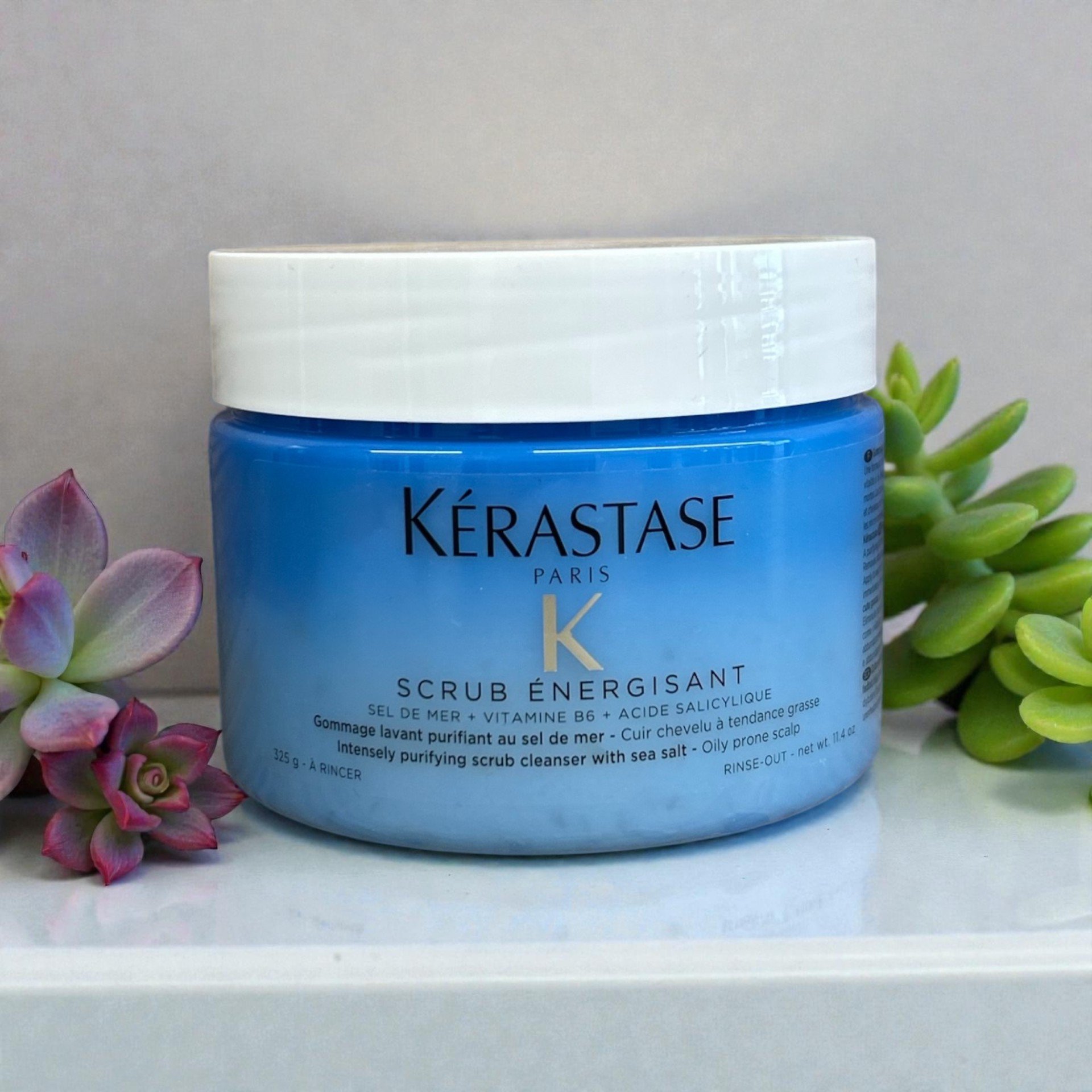 A blue jar of Kérastase Paris Scrub Énergisant hair scrub with sea salt, surrounded by pink and green succulent plants on a white shelf.