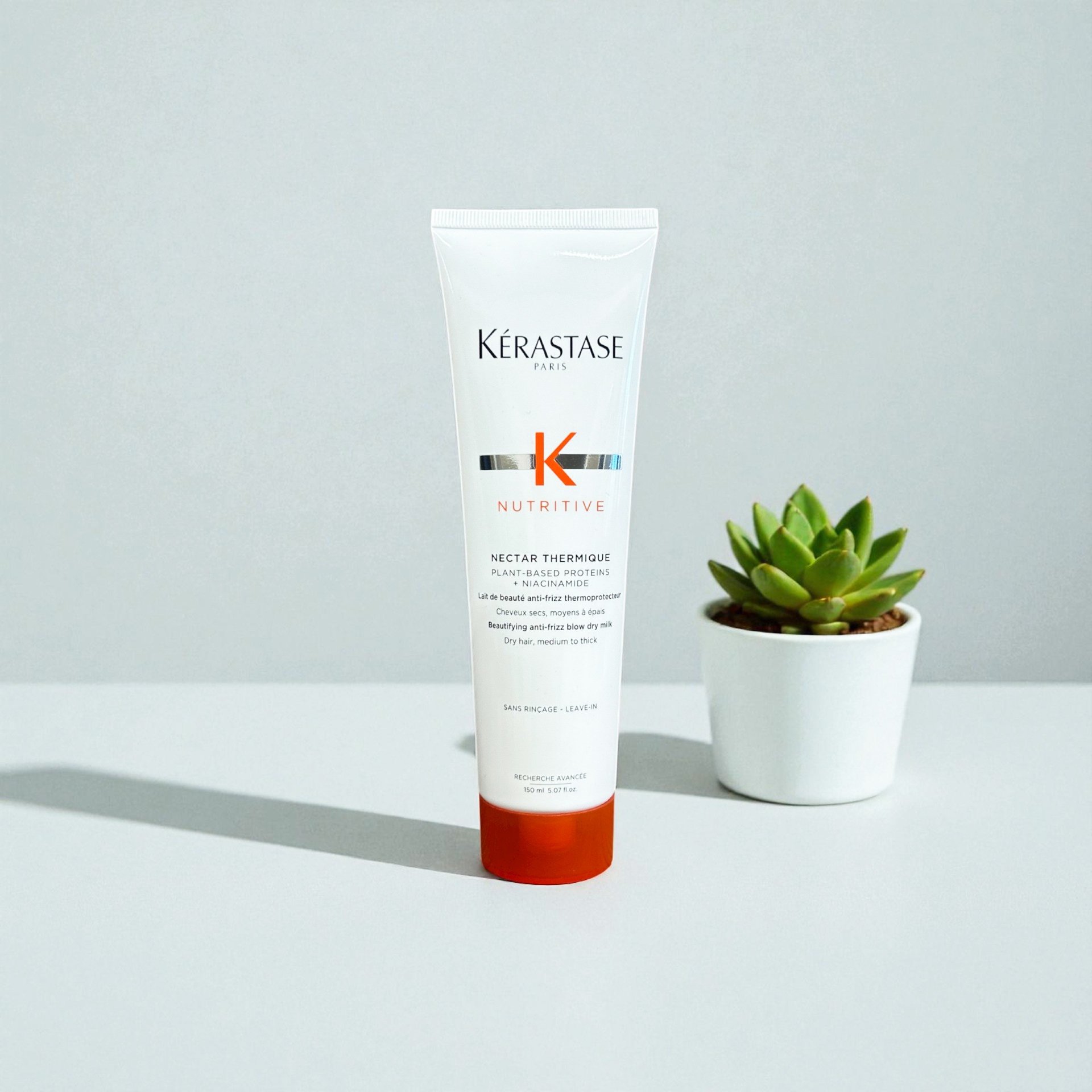 A tube of Kérastase Nutritive Nectar Thermique hair protective cream stands on a white surface beside a small potted succulent plant.