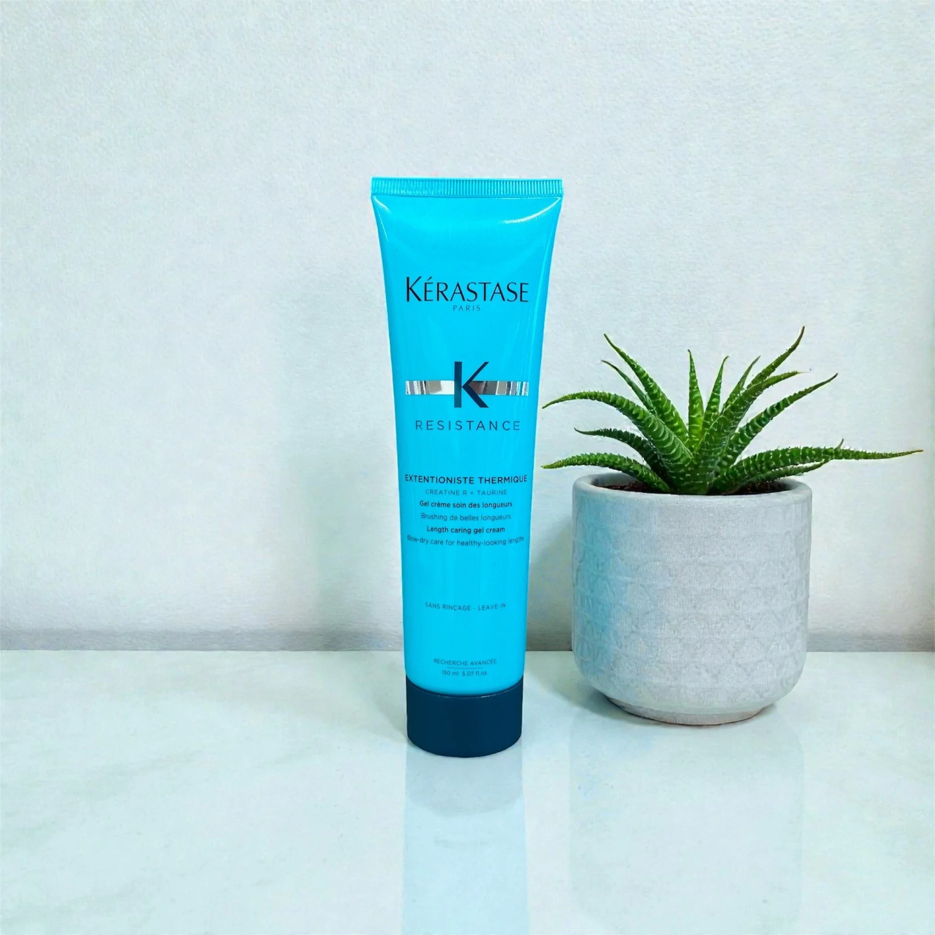 A tube of Kérastase Resistance Length Strengthening Gel Cream placed beside a potted succulent plant on a white surface against a plain background.