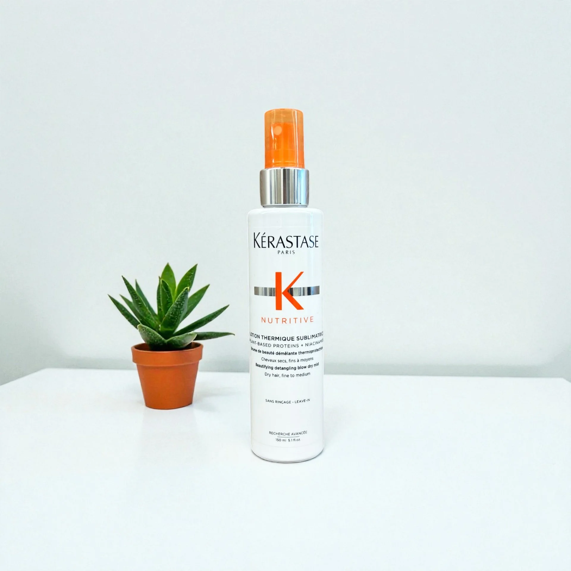 A white spray bottle of Kérastase Nutritive hair product placed on a white surface next to a small potted green succulent plant, against a plain light gray background.