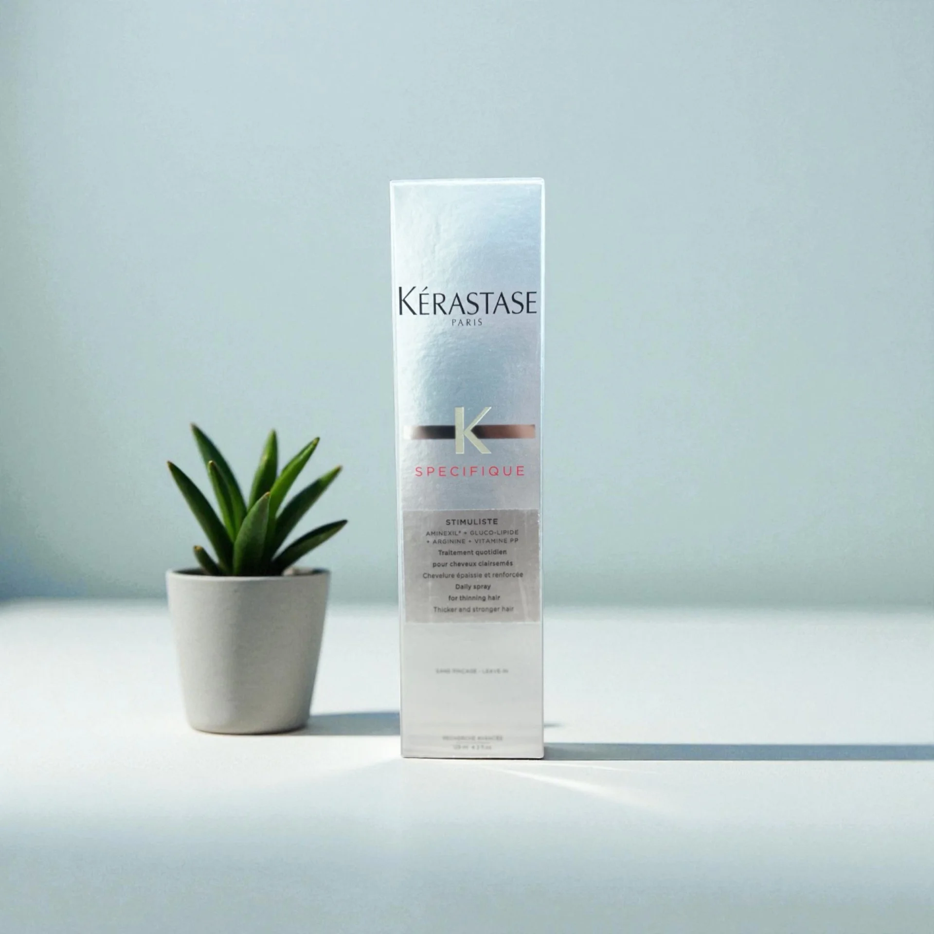 A silver box of Kérastase Paris hair product next to a small potted green succulent plant on a white surface.