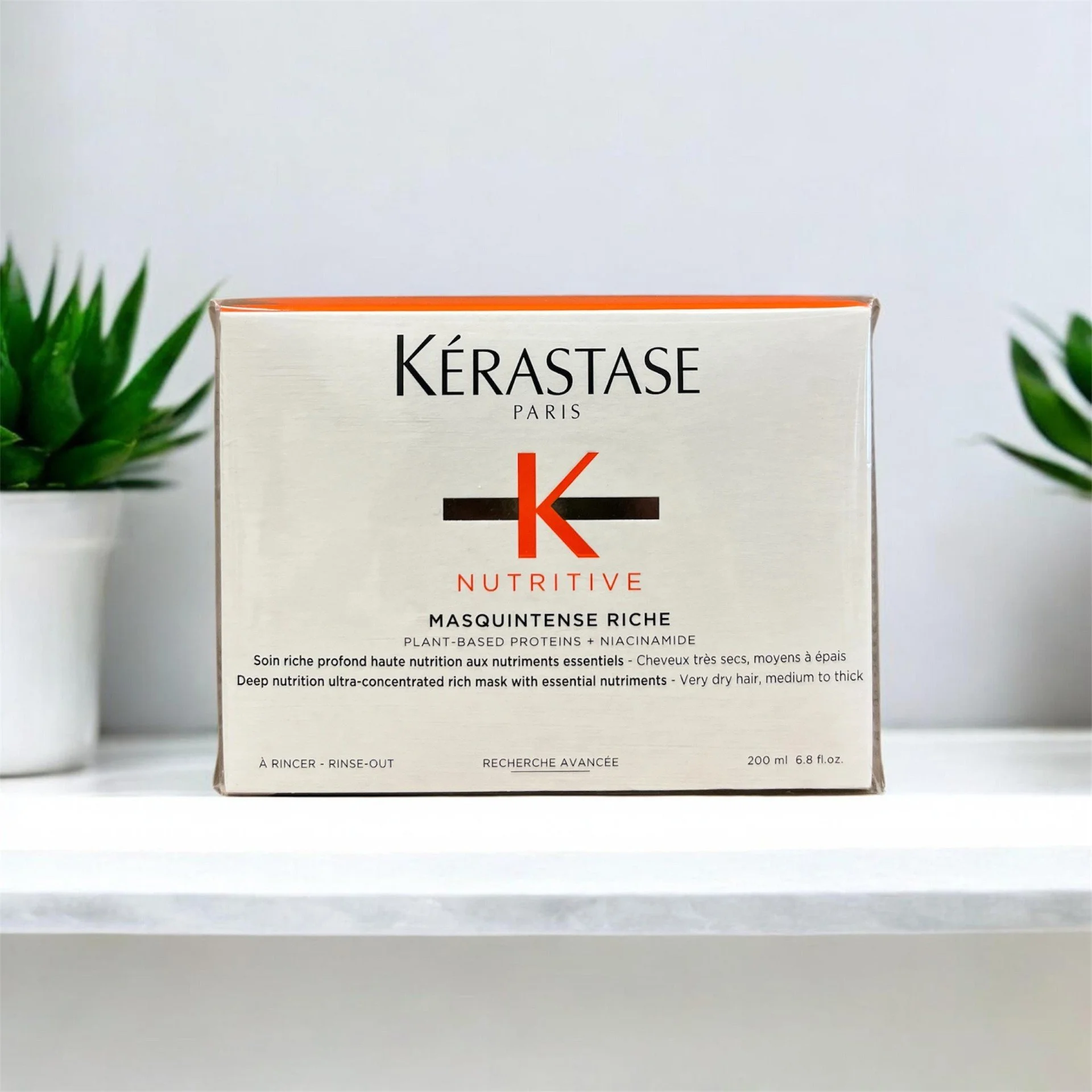 Kérastase Nutritive Masquintense Riche hair mask box on a white surface with green plants on either side in white pots.