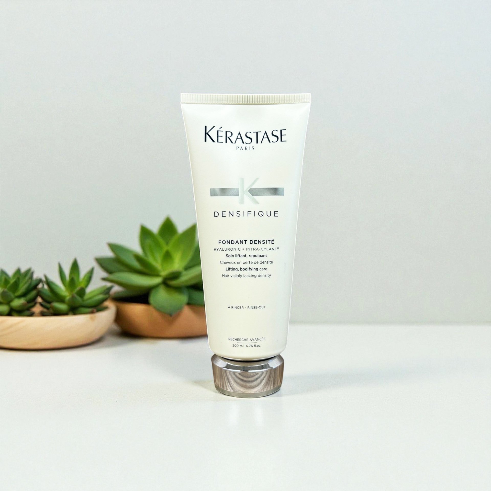 Kérastase Paris Densifique hair care product in a white tube with silver cap, placed on a white surface with green succulent plants in the background.