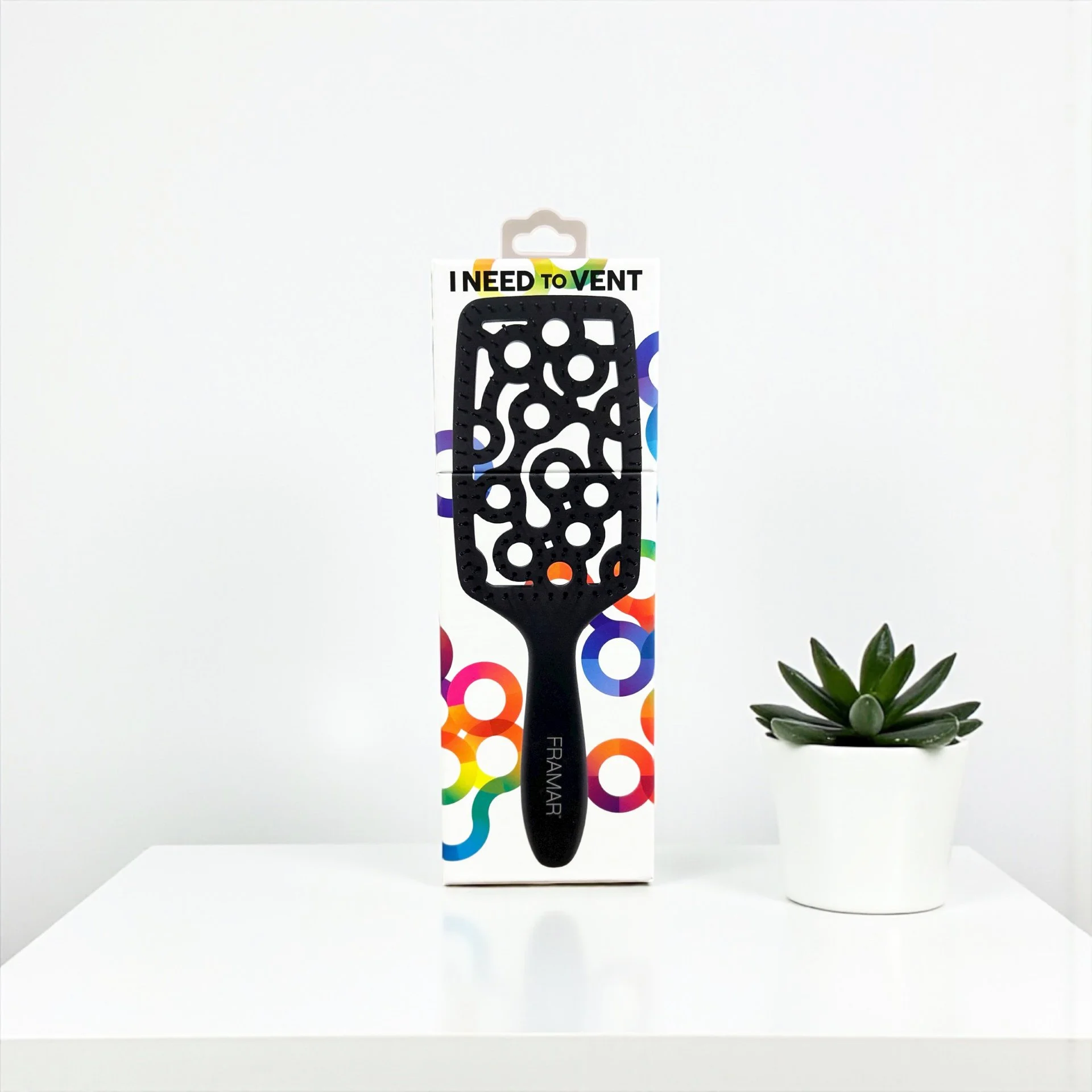 A black hair brush, labeled 'FRAMAR', standing upright in front of colorful packaging that says 'I NEED TO VENT', next to a small potted succulent plant on a white surface against a plain white background.