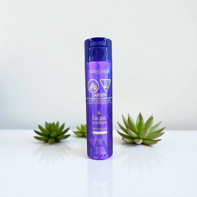 A purple can of Kérastase Laque Couture hair spray on a white surface, with green succulent plants in the background.