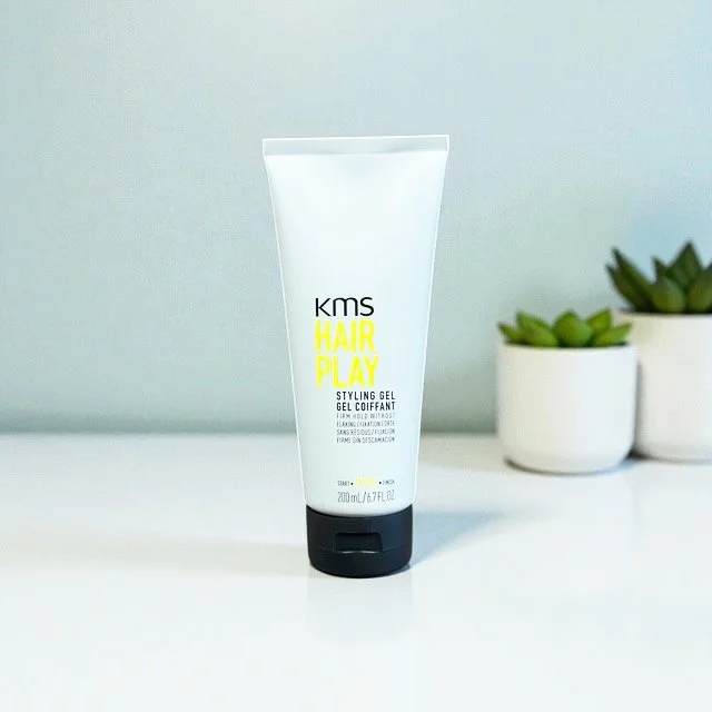 KMS Hair Play Styling Gel Coiffant in a white tube with black cap, placed on a white surface with potted succulent plants in the background.
