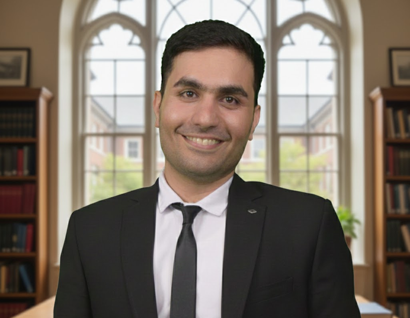 A smiling man in a black suit and tie inside a room with large windows and bookshelves.
