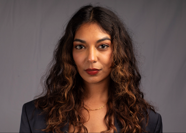A woman with curly brown hair and brown skin looking directly at the camera, wearing a dark blazer and a gold necklace against a plain gray background.