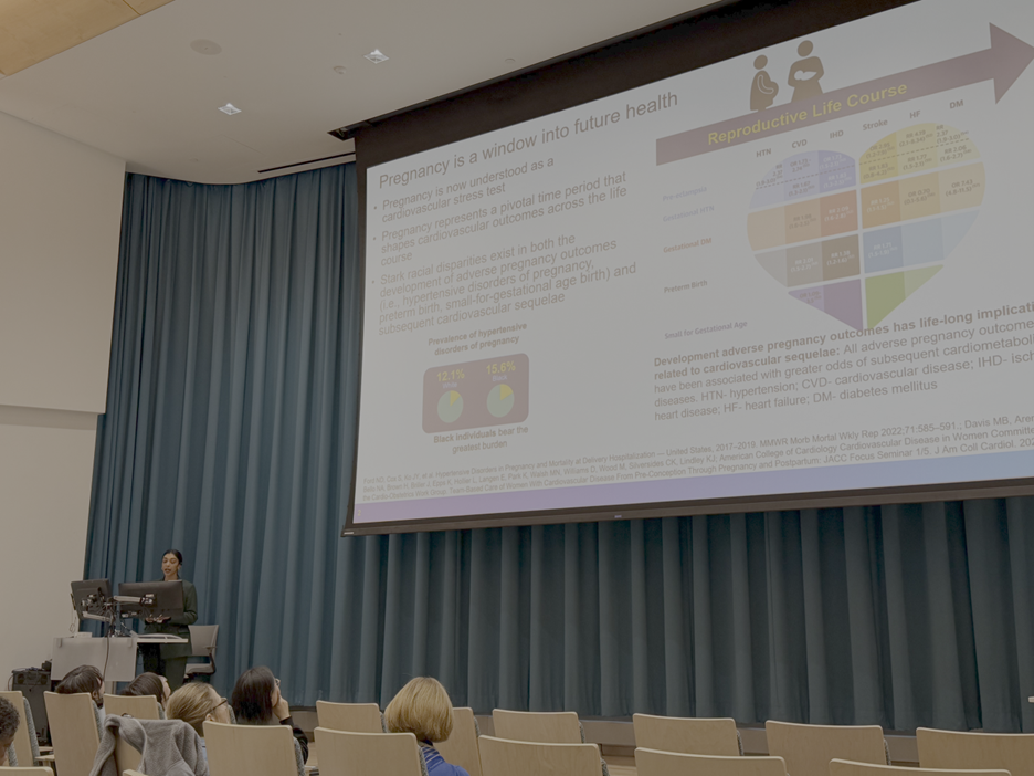 A woman presenter standing at a podium giving a presentation on pregnancy and future health, with a large slide projected on a blue curtain backdrop, in a conference room with seated audience members.