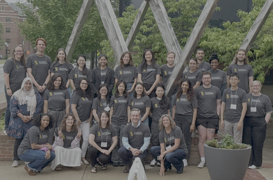 Group of diverse people wearing matching gray T-shirts with logos, standing outdoors in front of a modern metal sculpture and trees.