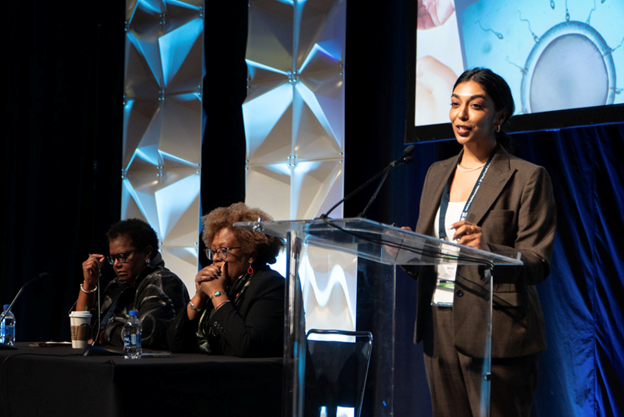 A woman giving a presentation at a conference, with two women seated at the table beside her, and a large screen displaying graphics behind them.