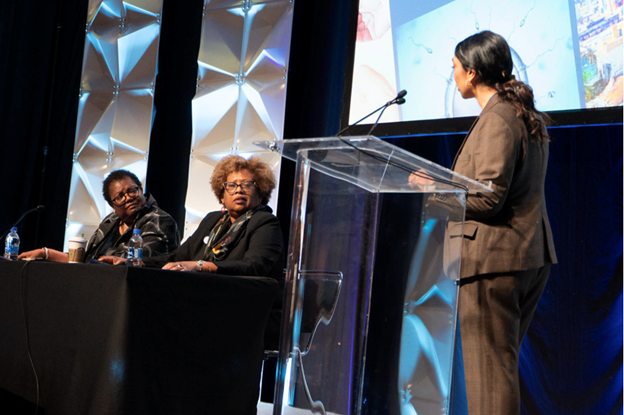 A woman speaking at a podium during a panel discussion, with two women sitting at a table on stage and a large screen in the background showing visuals, all in a conference setting.
