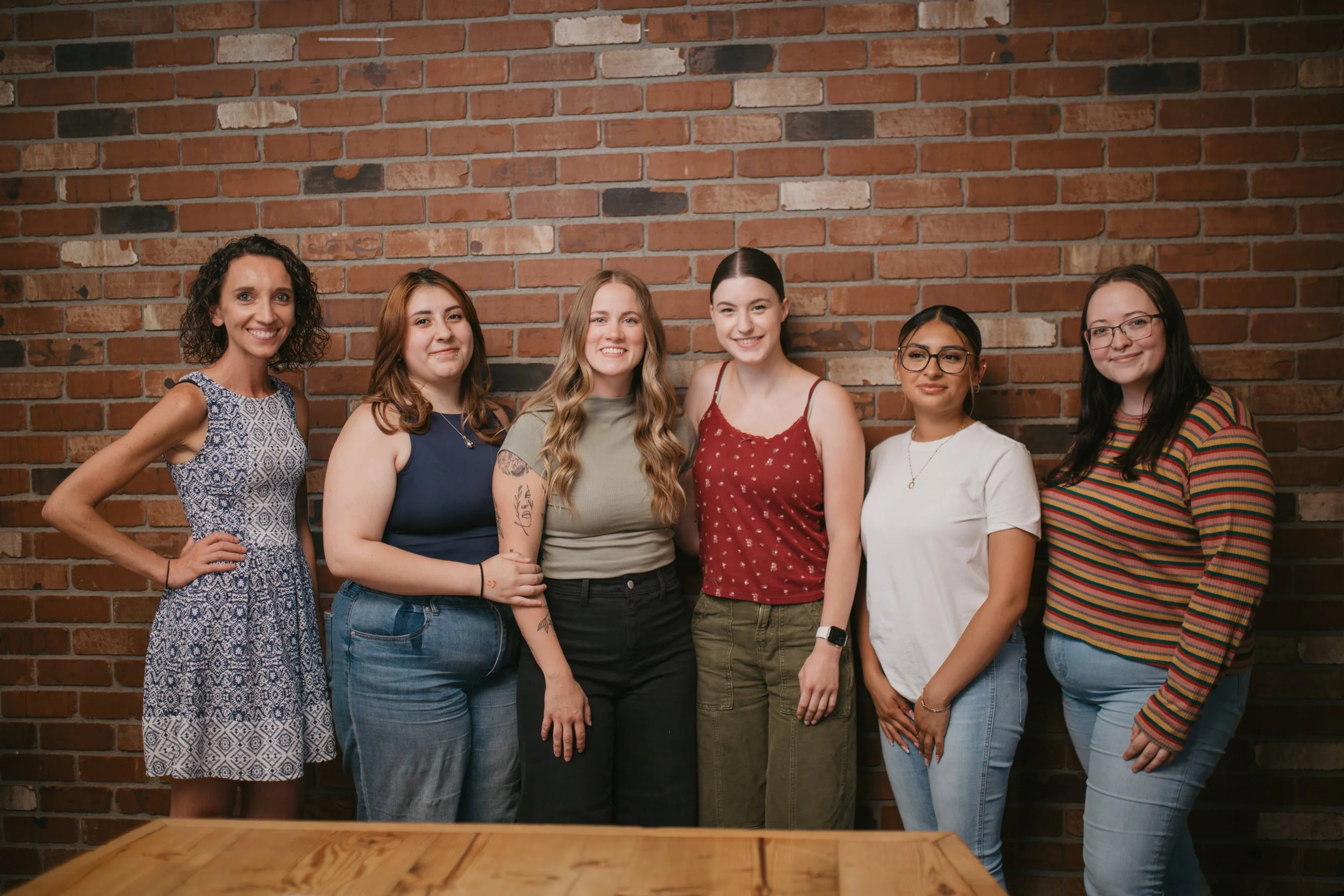 Group of six women standing indoors in front of a brick wall, smiling at the camera.