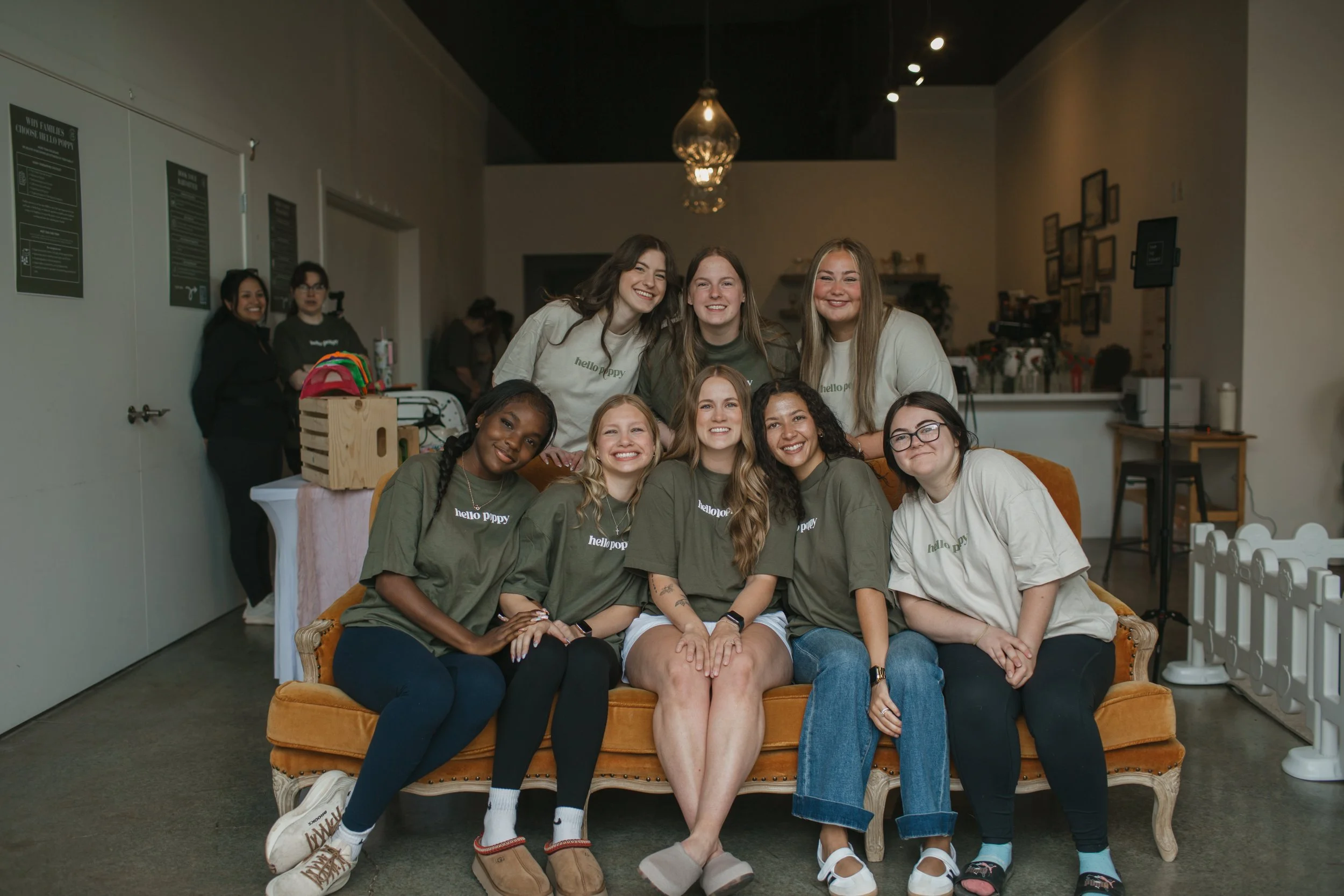 Group of nine young women sitting and standing on a vintage orange sofa in an indoor space, smiling for the photo. Some are wearing matching dark green shirts with