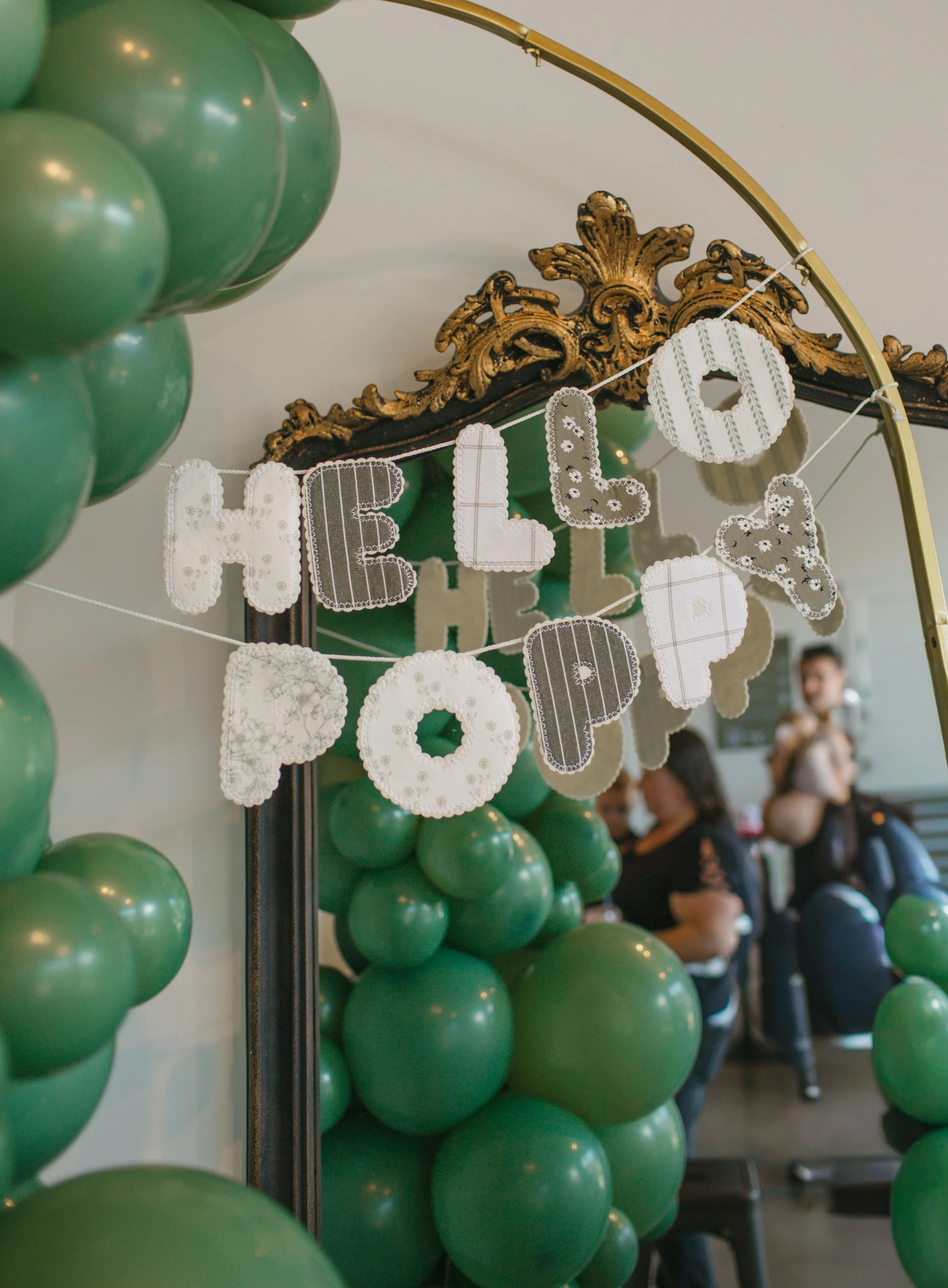 Green balloons arranged around a mirror decorated with a white and pastel-colored banner that reads 'HELLO PAPA'. In the background, people are seated and taking photos.