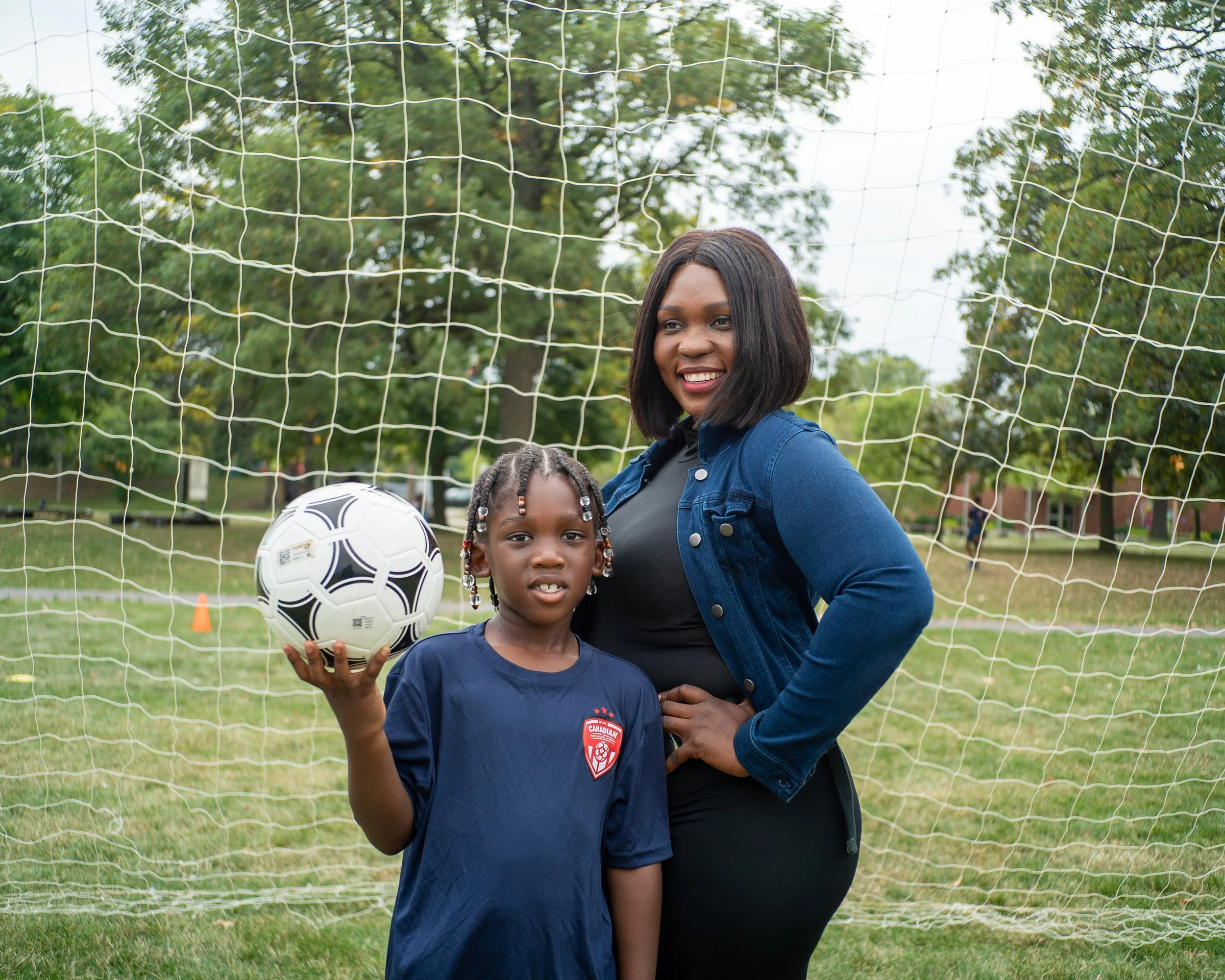 A woman and a young girl standing in front of a soccer goal net in a park, with trees in the background. The girl is holding a soccer ball and wearing a navy blue sports shirt with a red and white badge. The woman has dark hair, is smiling, and is wearing a blue jacket over a black shirt.