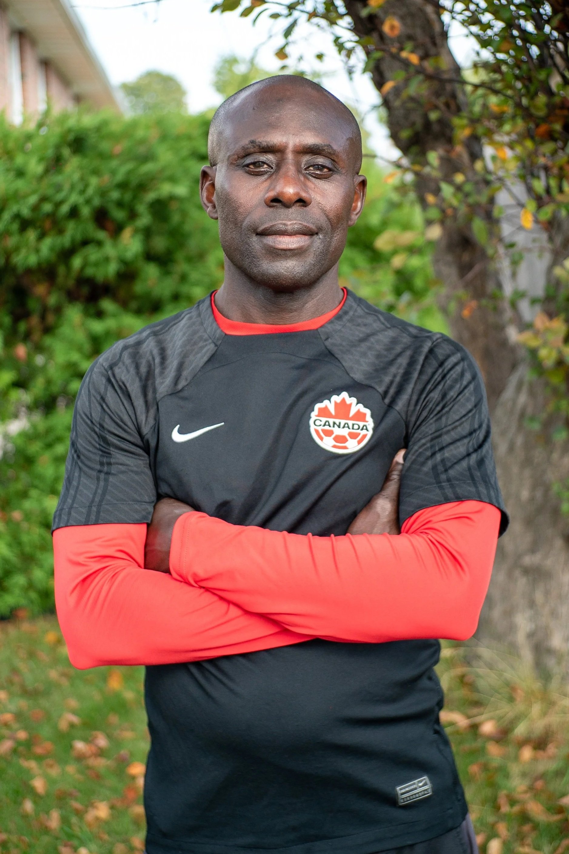 A man with dark skin and short hair standing outdoors on a sports field, wearing a red jacket with a white zipper and the word "Canada" on it, crossing his arms and looking confidently at the camera.