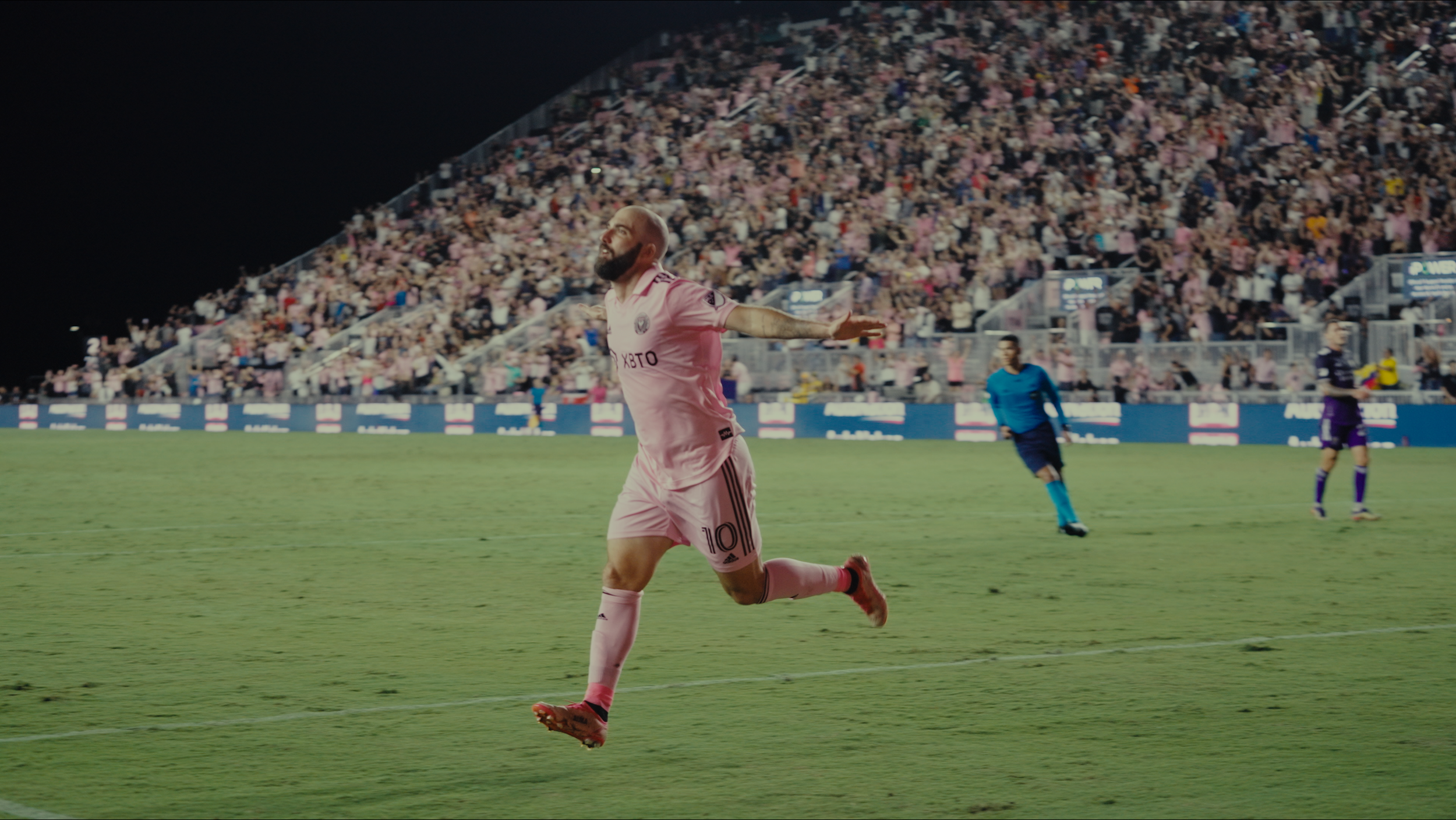 Soccer player in pink uniform celebrating on the field with arms outstretched, crowd in the stands behind him, nighttime stadium setting.