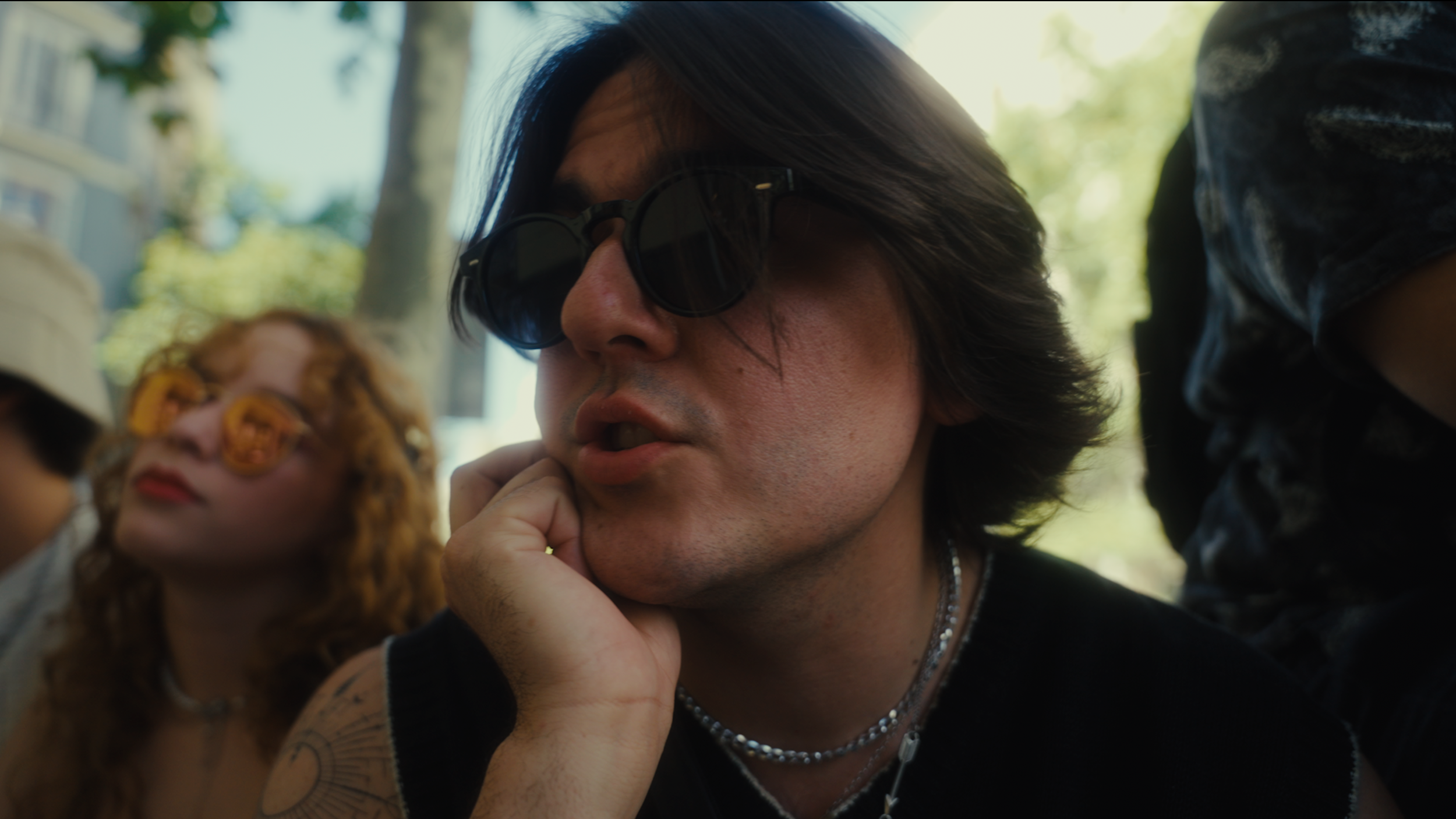 Close-up of a man with long dark hair wearing sunglasses, resting his chin on his hand, during daytime outdoors.