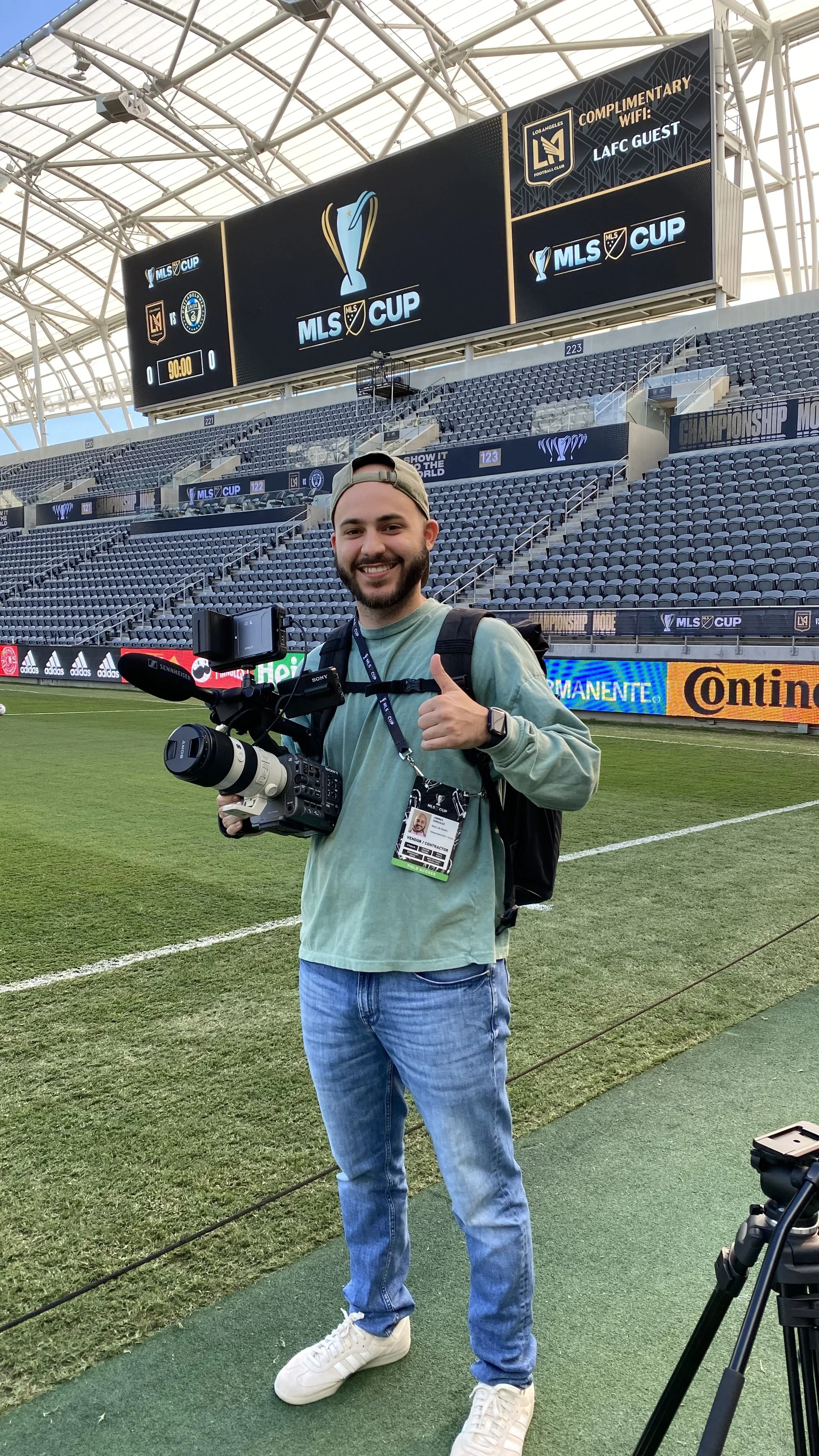 A smiling man holding a professional camera with a microphone attached, standing on a soccer field in an empty stadium, with a large screen showing MLS Cup and Los Angeles Football Club advertisements in the background.