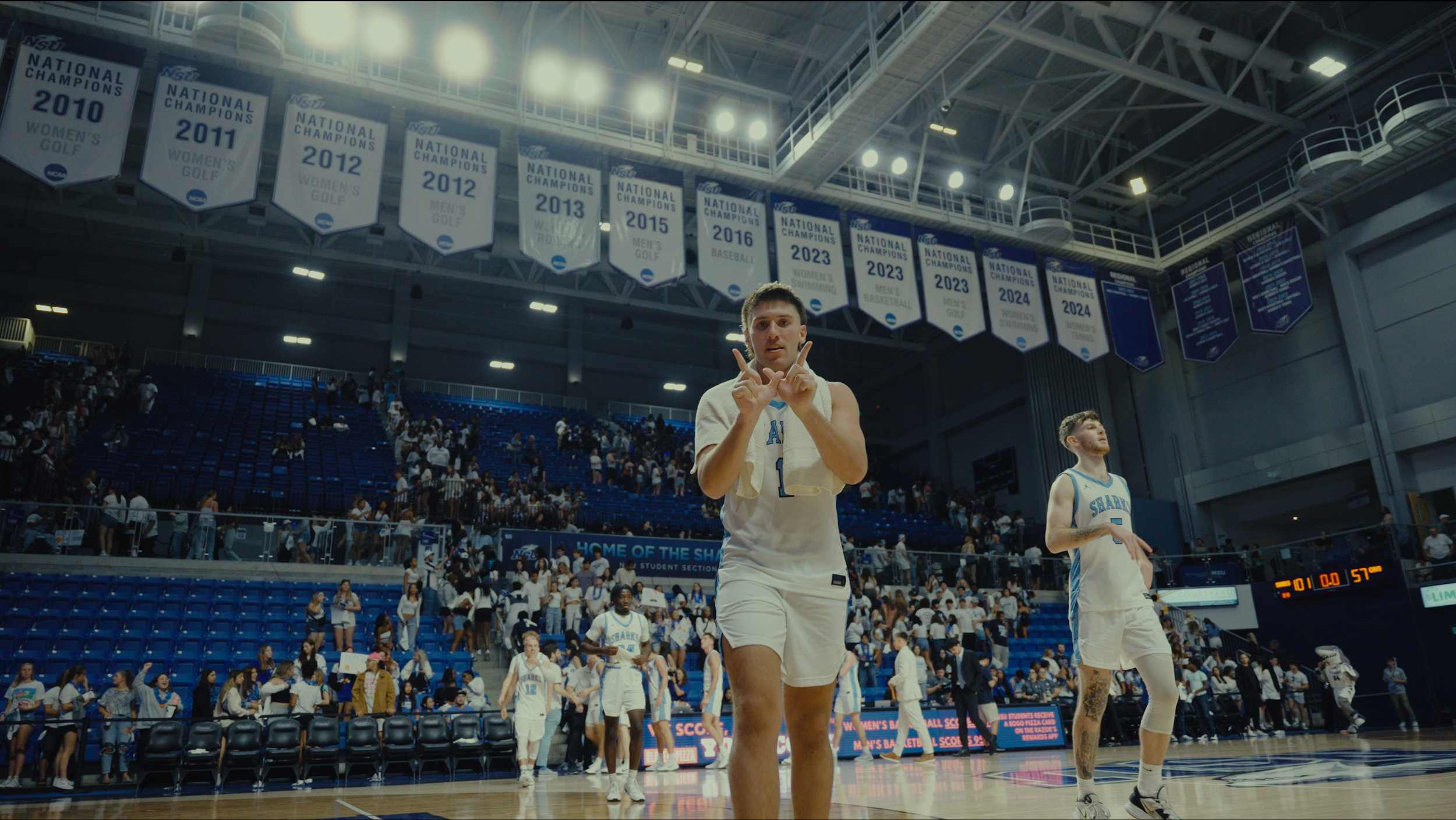 A basketball player with a towel around his neck, standing on the court with both hands making peace signs, surrounded by teammates and fans in a large indoor arena with banners hanging from the ceiling.