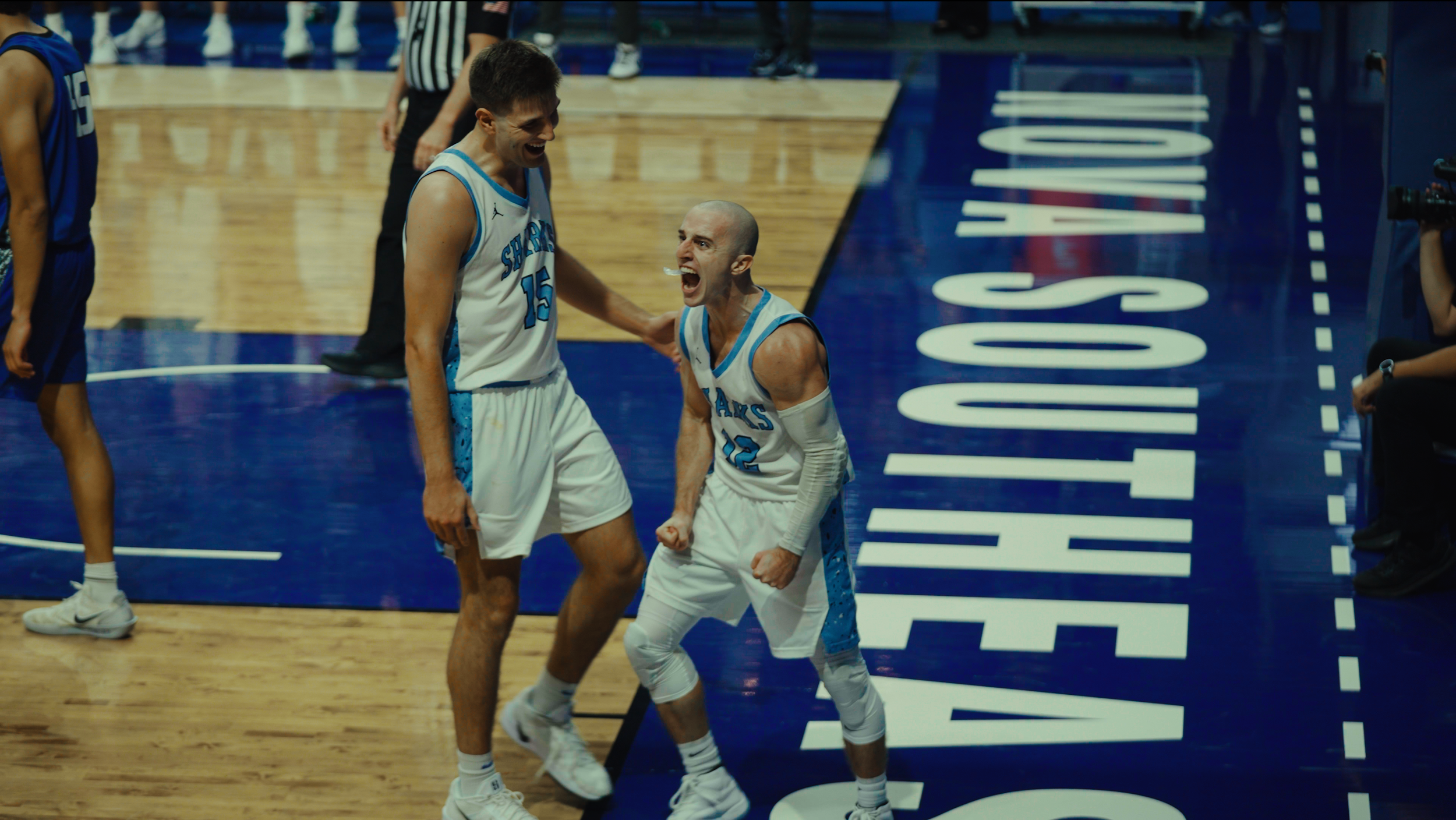 Two basketball players celebrating on the court, one is taller and the other shorter, both wearing white and blue uniforms with the numbers 15 and 12, with the word 'SHARKS' on their jerseys. They are expressing excitement and joy.