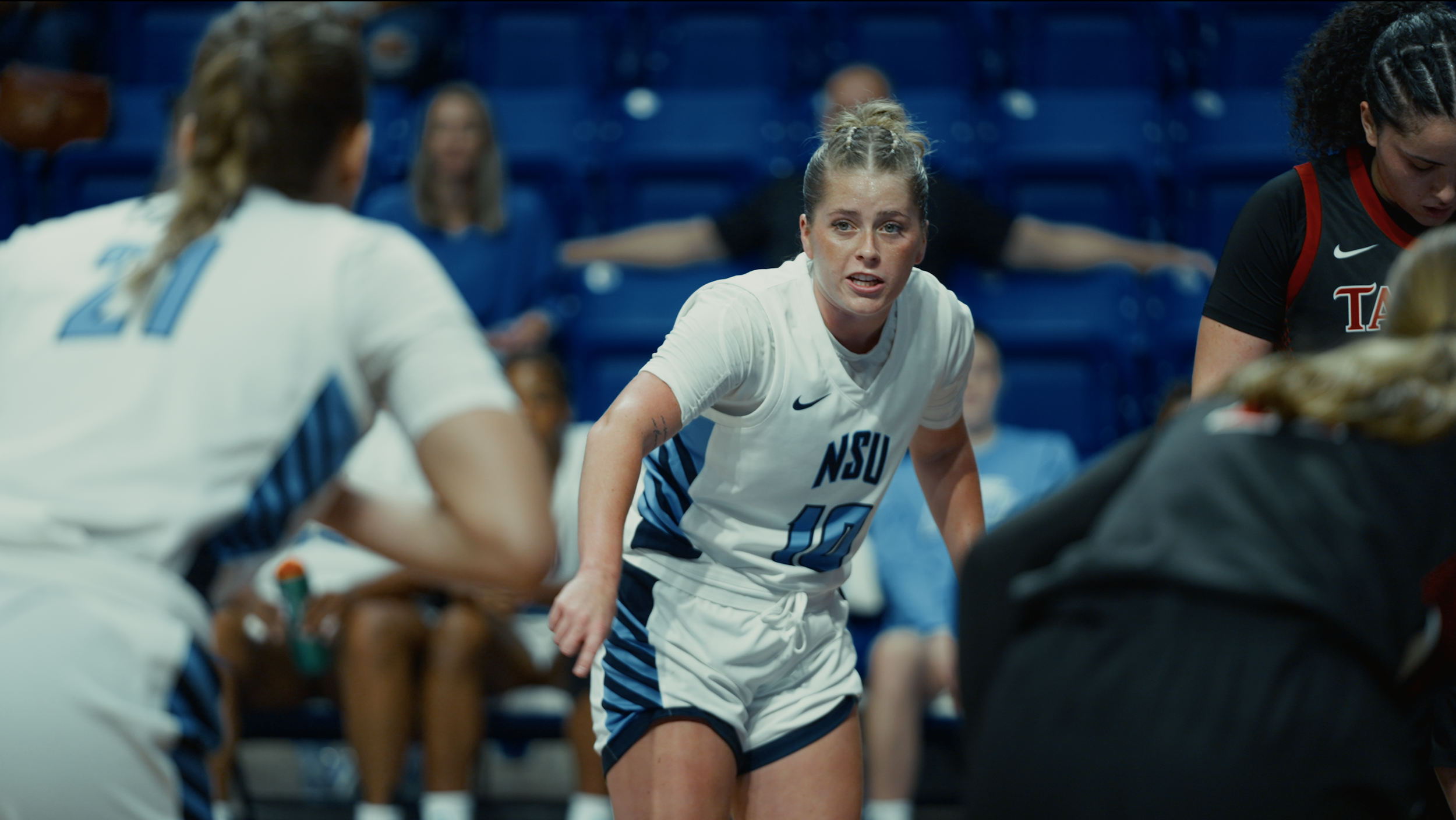 Female basketball players in a game, with one player in white jersey with NSU logo, leaning forward and talking to teammates