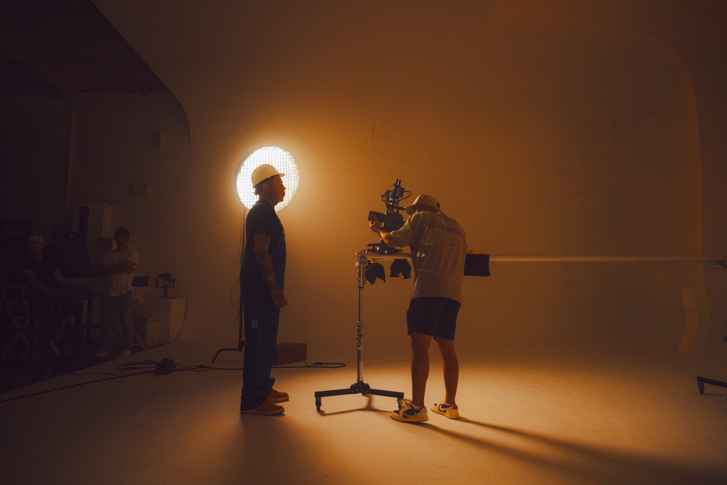 Filming scene with two crew members, one using a camera and the other in a white shirt and hard hat, in a dimly lit studio with a large circular light behind.