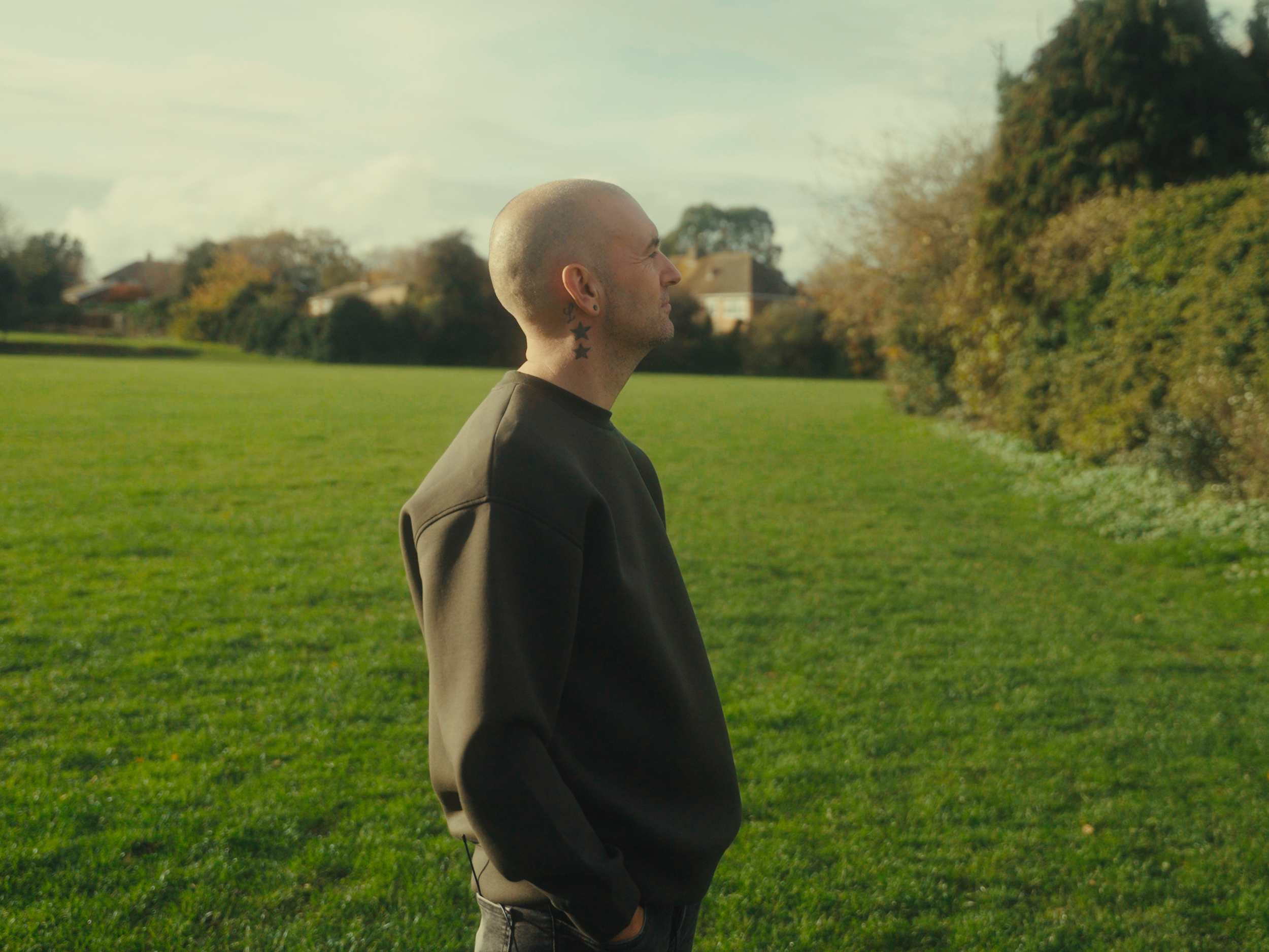 A man with a shaved head and star tattoos on his neck, standing outdoors in a green field with trees and houses in the background, during daytime.