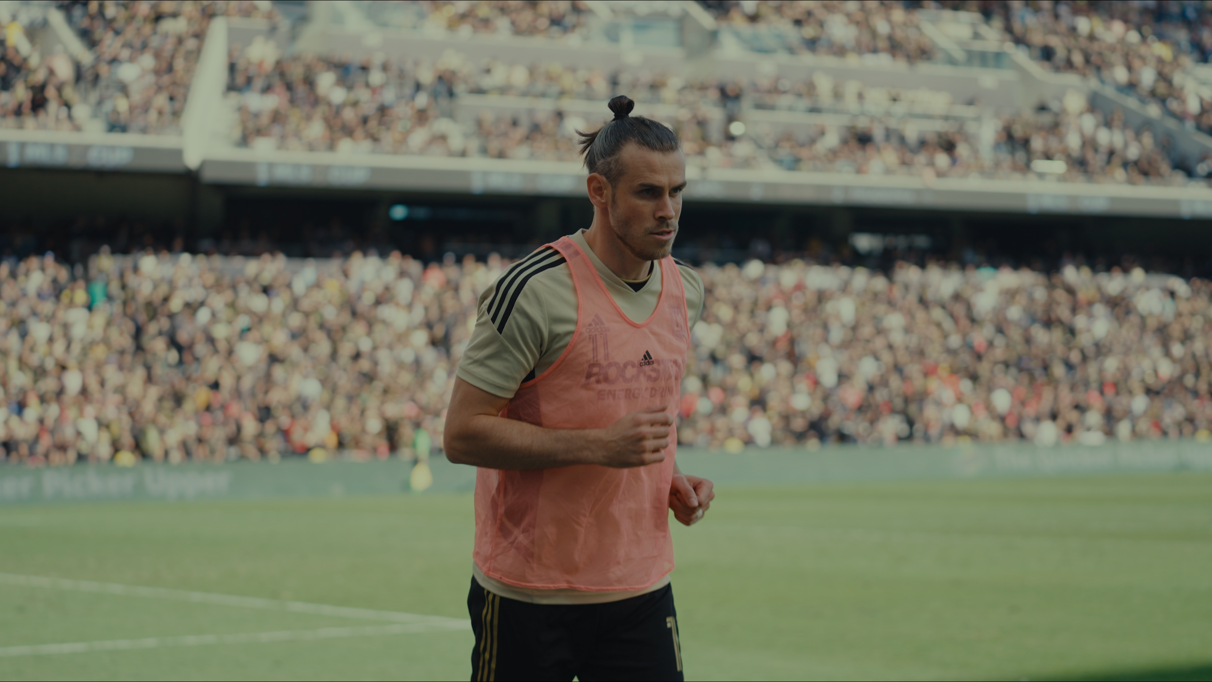 A male soccer player with long hair tied back in a bun, wearing a peach-colored training vest over a beige Adidas jersey, jogging on a soccer field during a match with a large crowd in the stadium background.