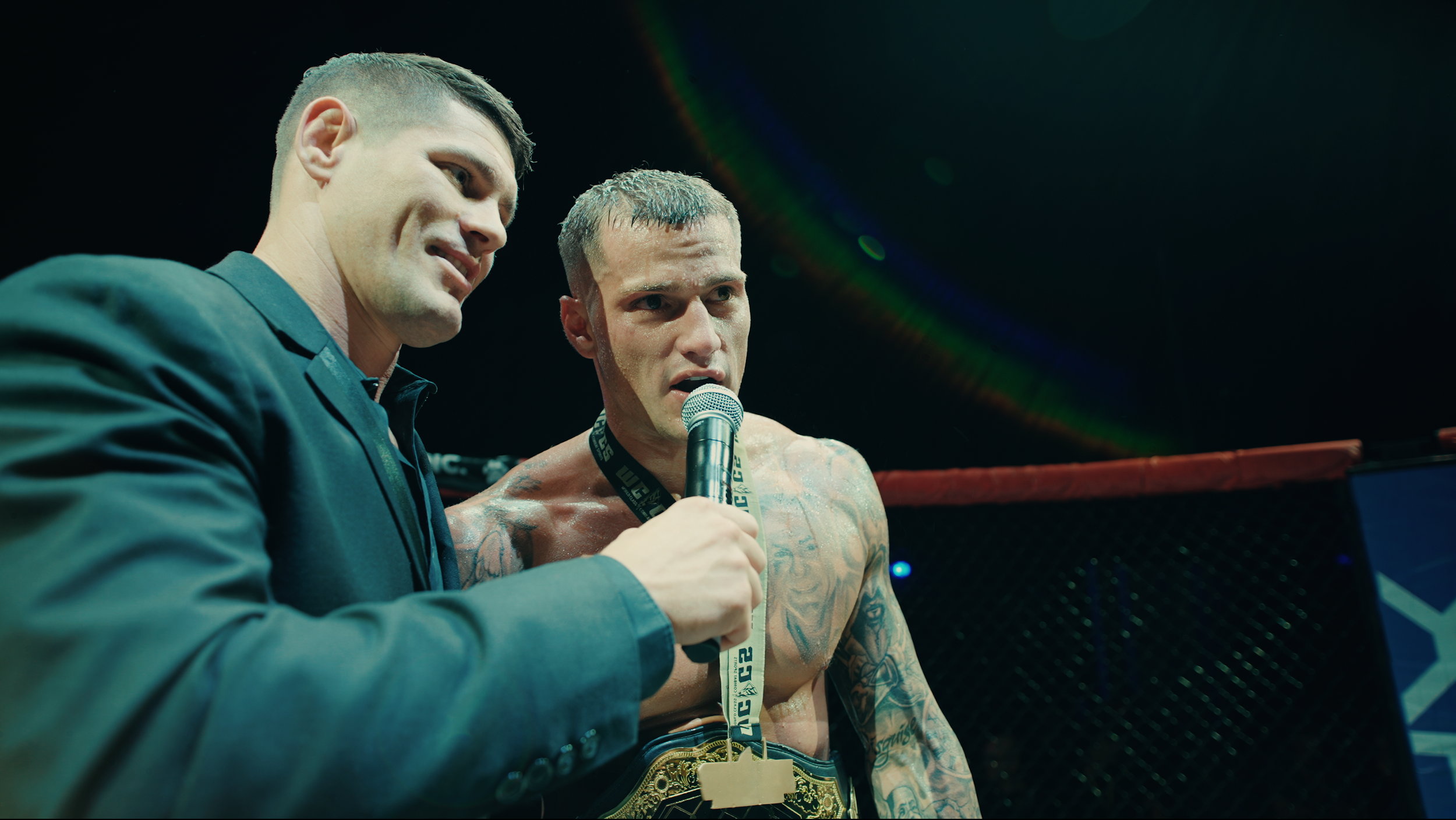 A male MMA fighter with tattoos on his chest and arm being interviewed in the octagon after a match, wearing a championship belt, while a man in a suit holds a microphone in front of him.