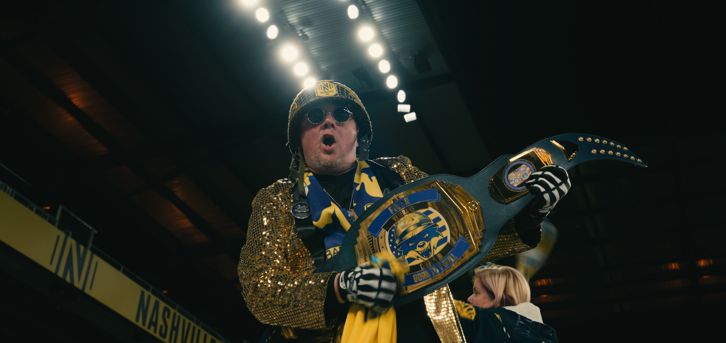 A man wearing a gold sequined jacket, sunglasses, and a yellow hat is holding a guitar-shaped championship belt. He appears to be cheering or singing at an indoor event with bright overhead lights.