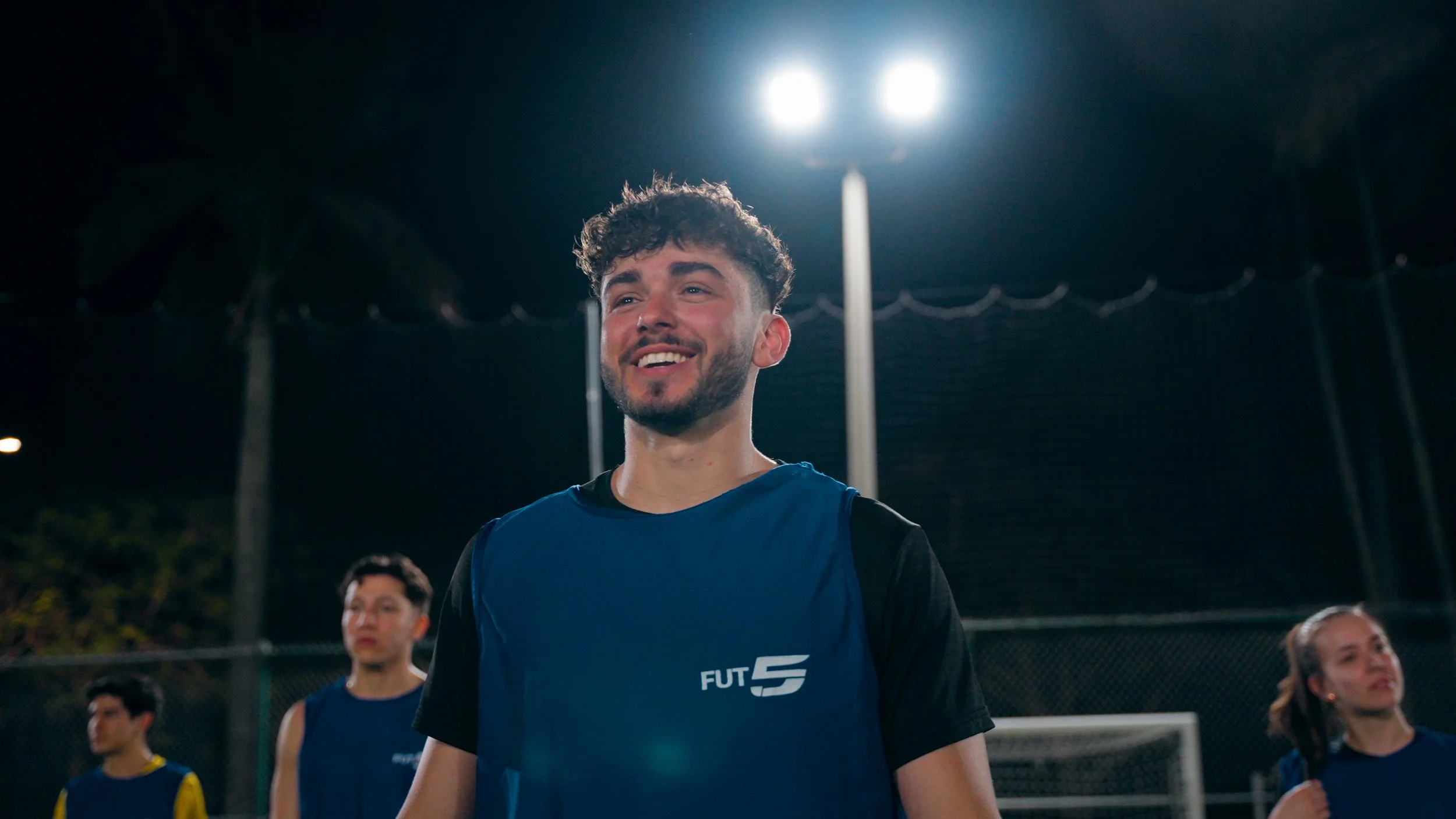 Young man smiling on a dark outdoor sports field at night, wearing a blue sports vest with the logo FUT 5, with other players in the background.