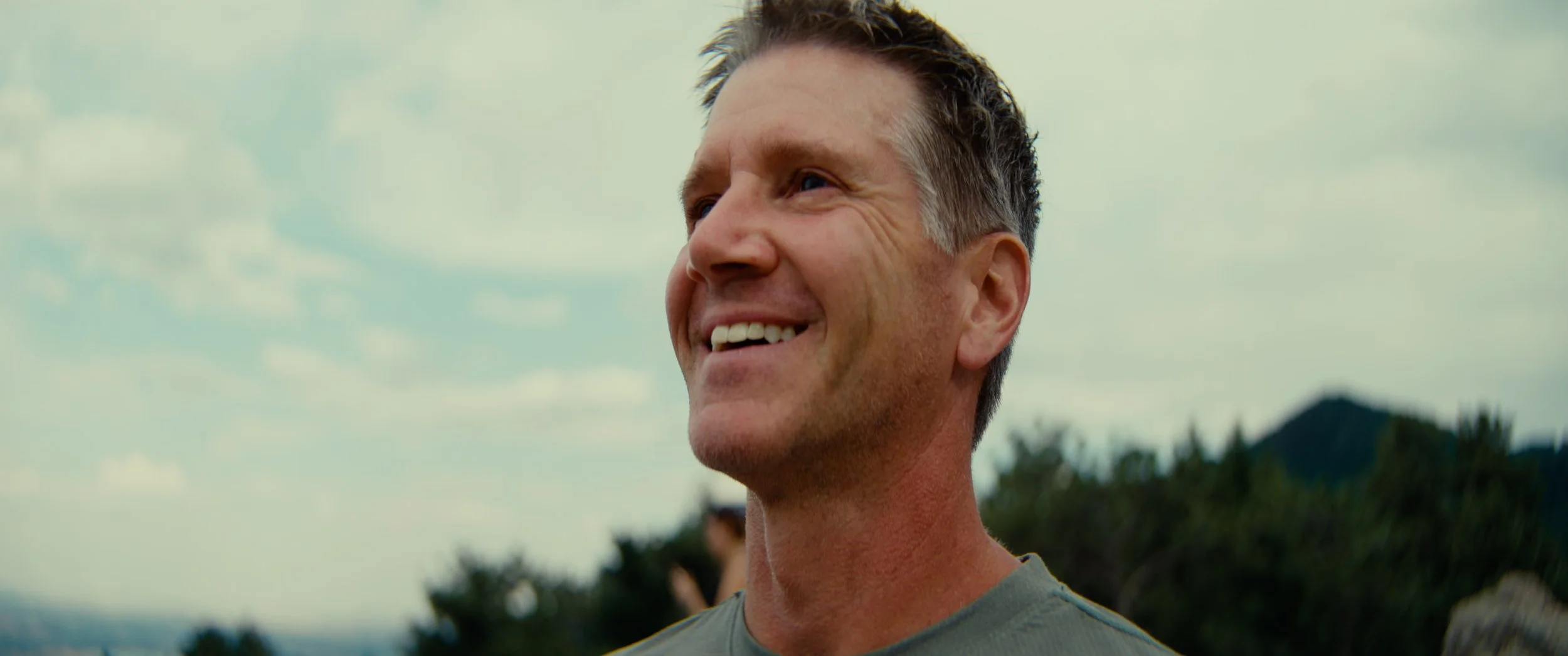 A middle-aged man with short, dark and gray hair smiling outdoors during daytime with a blurred background of trees and a cloudy sky.