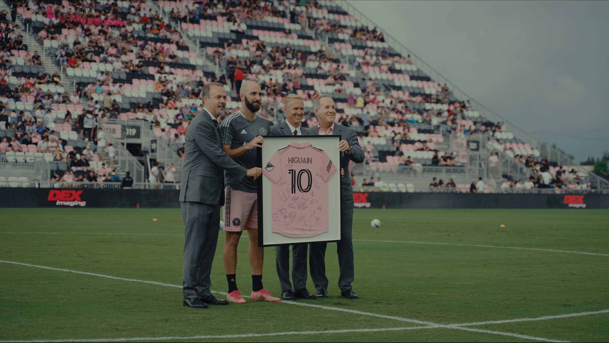 Four men standing on a soccer field, holding a framed pink jersey with the number 10 and autographs, with spectators in the stands behind them.