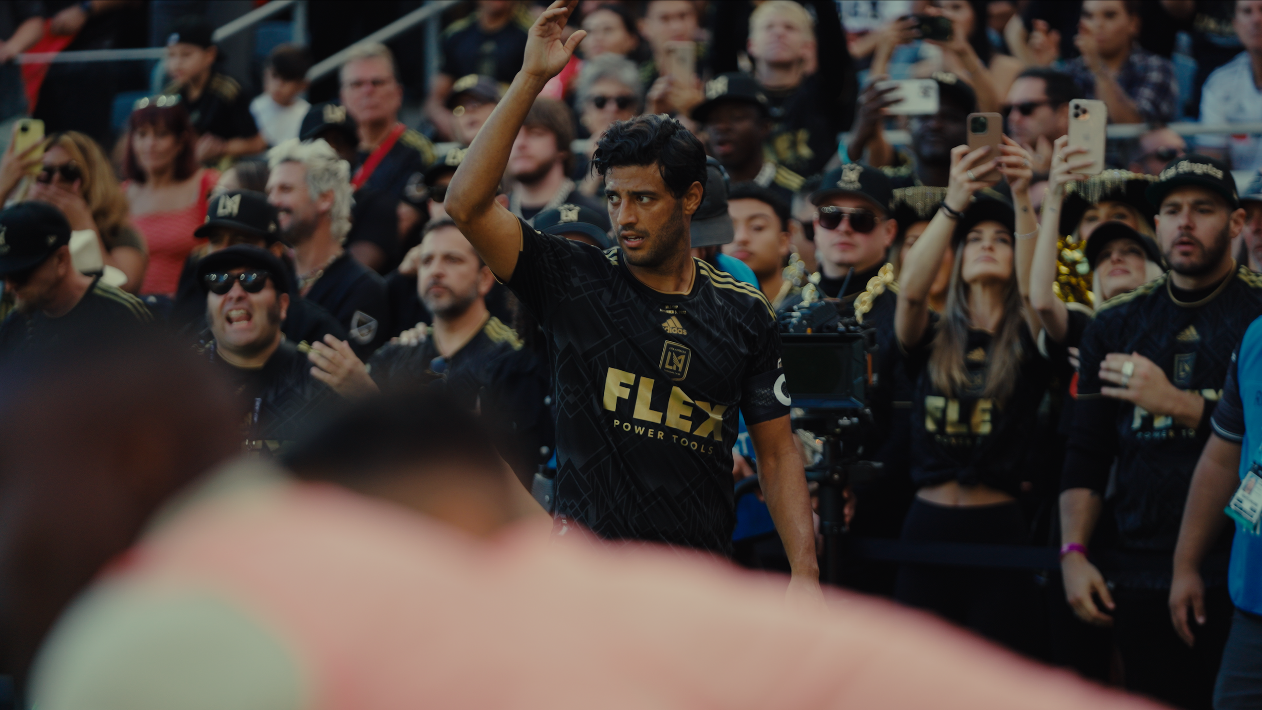 A man wearing a black sports jersey with gold accents and the Atlanta United logo is surrounded by a crowd of fans, some of whom are holding up phones, at a sports stadium or event.