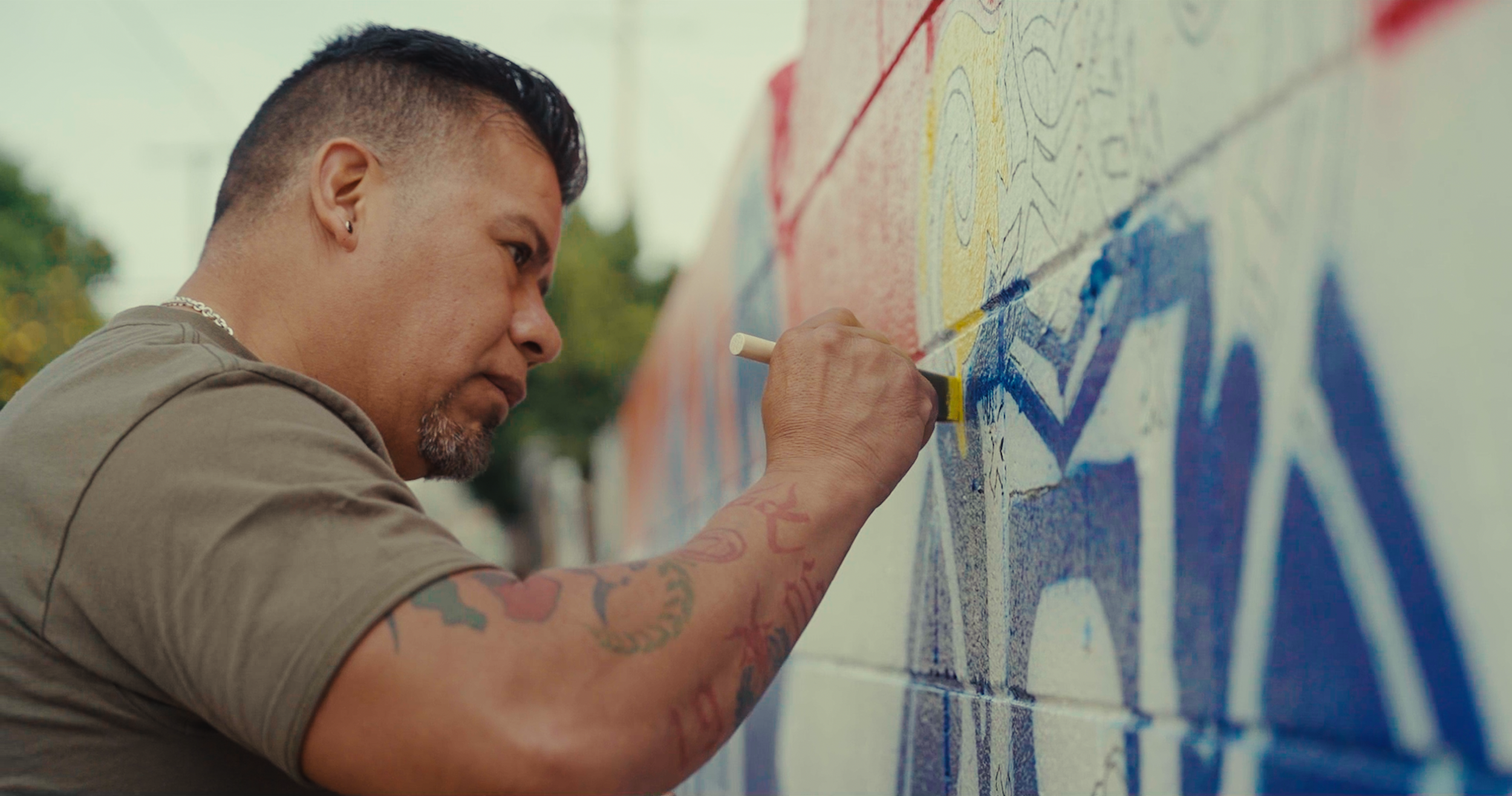 A man with tattoos on his arm is painting colorful graffiti on a wall with spray paint.