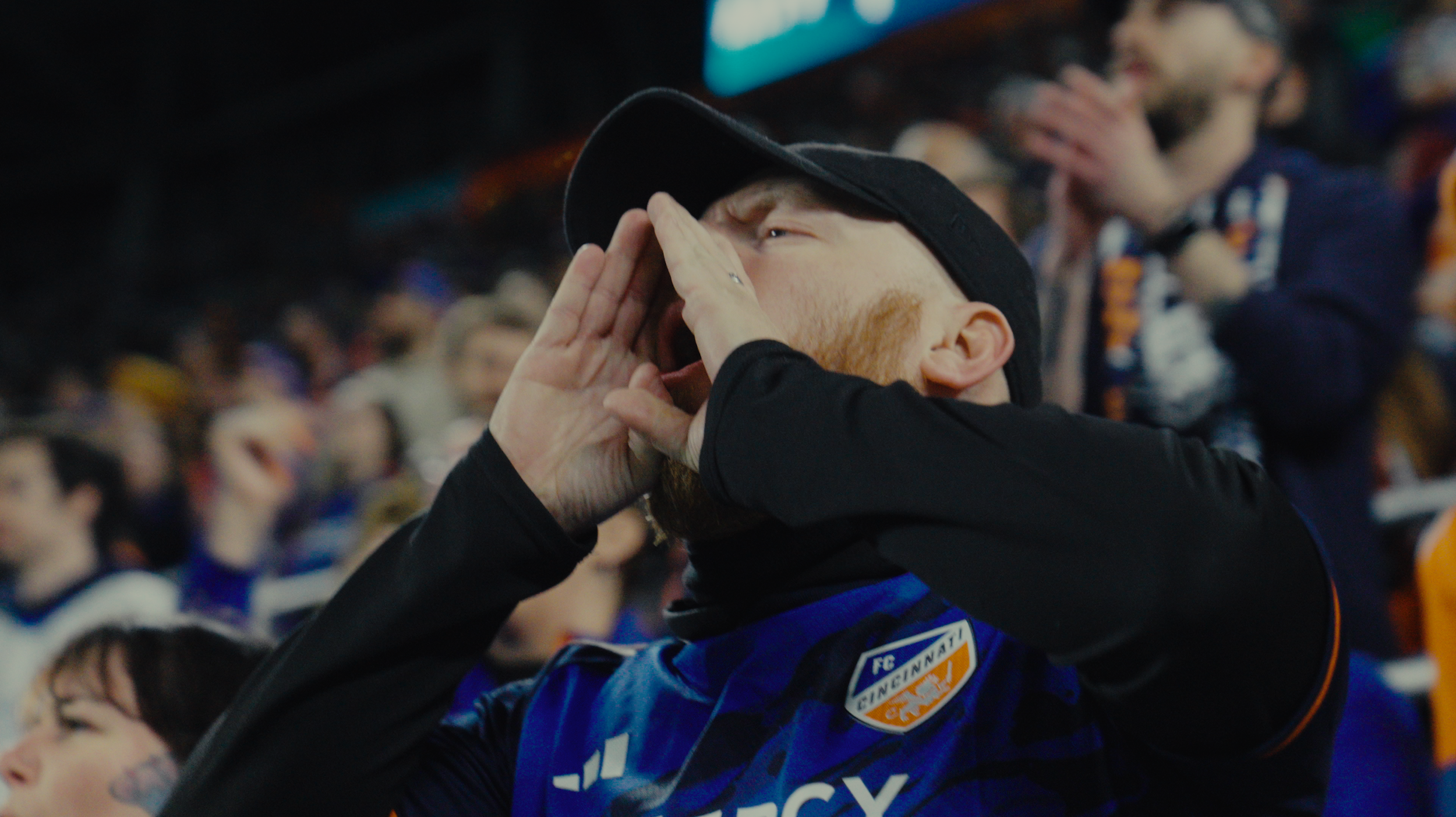 A man with a beard calling out or cheering at a sporting event, wearing a black cap and a blue sports jersey with the FC Cincinnati logo.