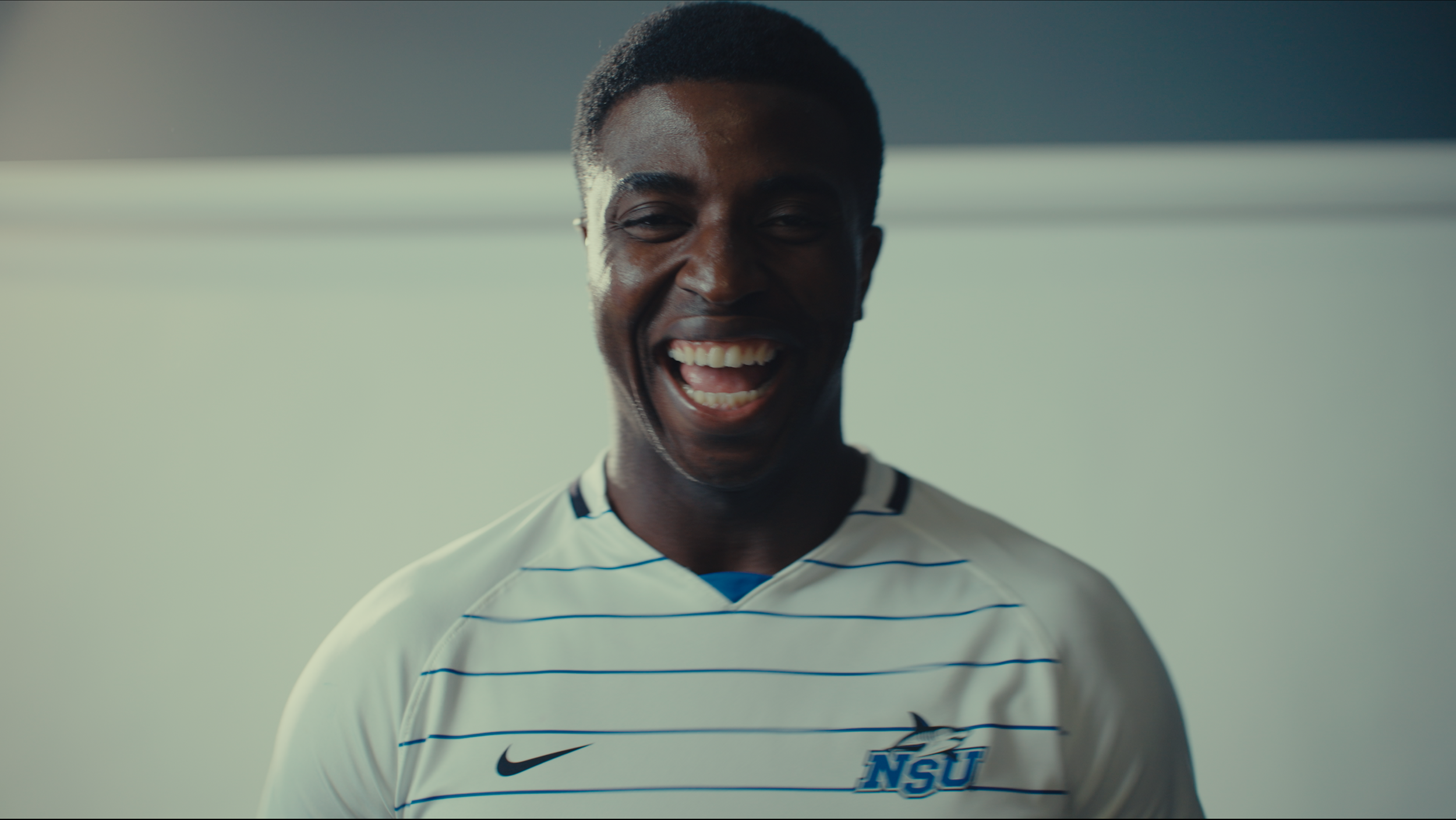 A young man smiling and laughing, wearing a white sports jersey with blue stripes and the 'NSU' logo, standing against a plain background.