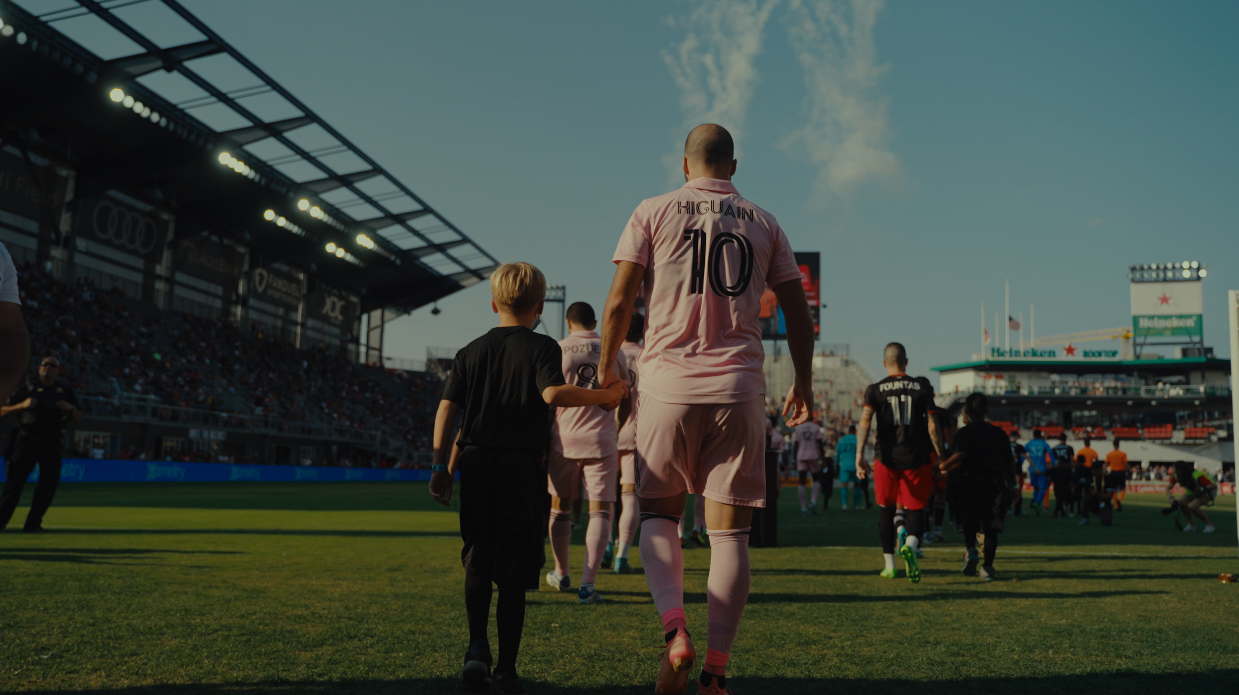 Soccer players and children walking onto a field before a game at a stadium, with a crowd in the stands and blue sky overhead.