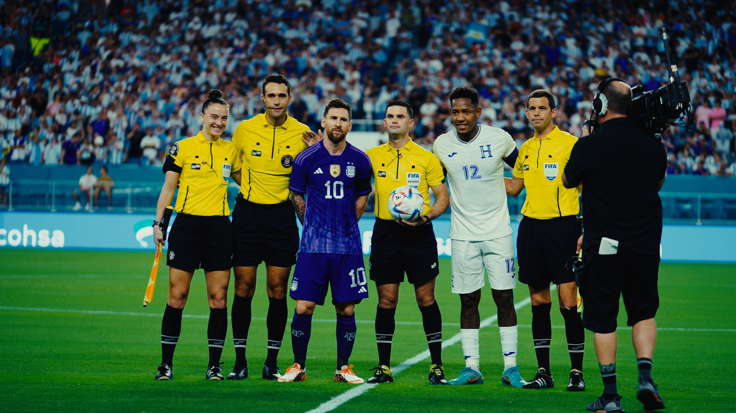 Soccer players and referees standing on the field during a match, with a cameraman filming them. The background shows a large crowd in the stands.